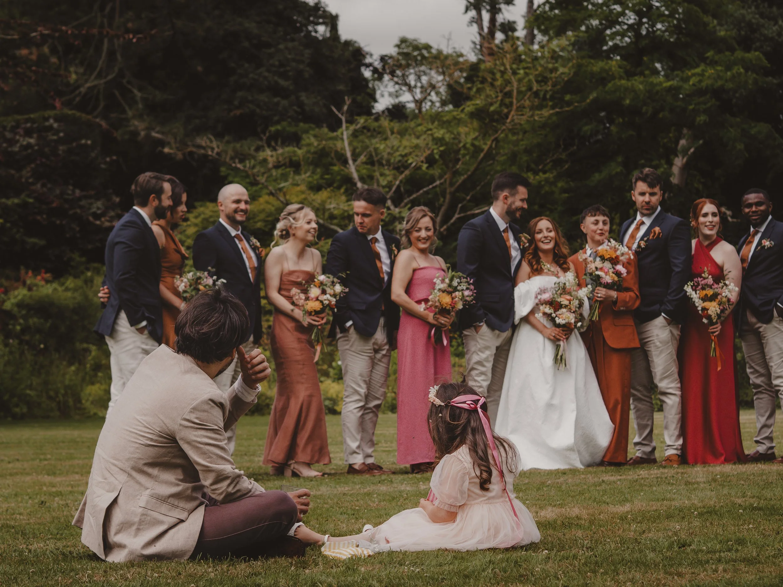 Wedding Party at Whatton House Tipis dressed in formal, colourful attire pose for a photo outdoors, with a dad and his young daughter sitting in the foreground, on a grassy field with trees in the background.