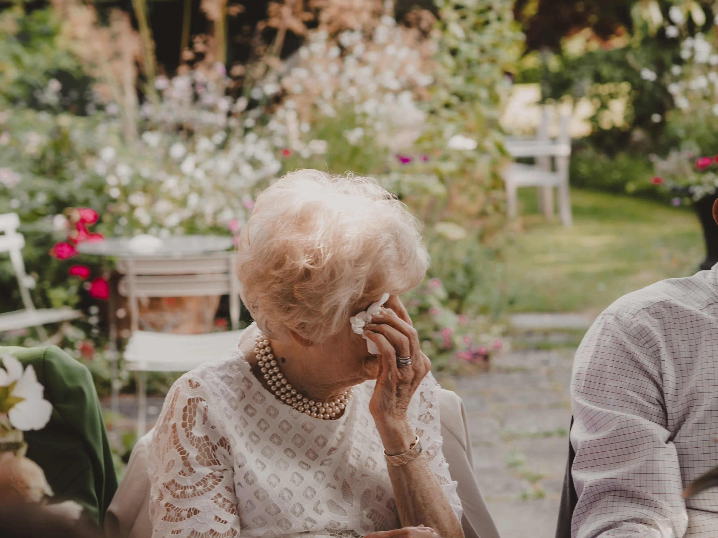 An elderly woman with white hair wiping her eyes with a tissue during an intimate wedding, surrounded by flowers and greenery.