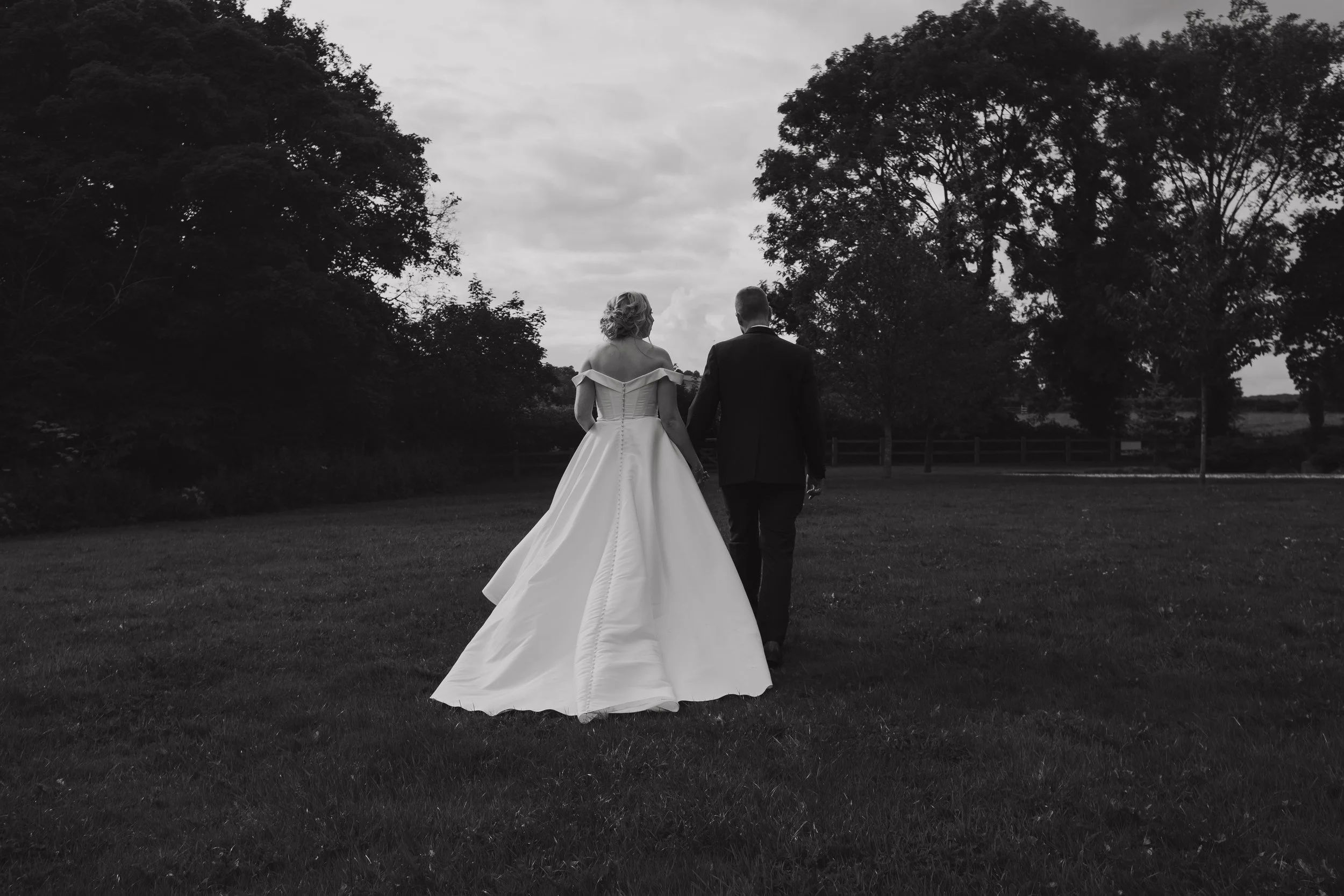 A bride and groom walking hand in hand outdoors, seen from behind, with trees and a cloudy sky in the background.