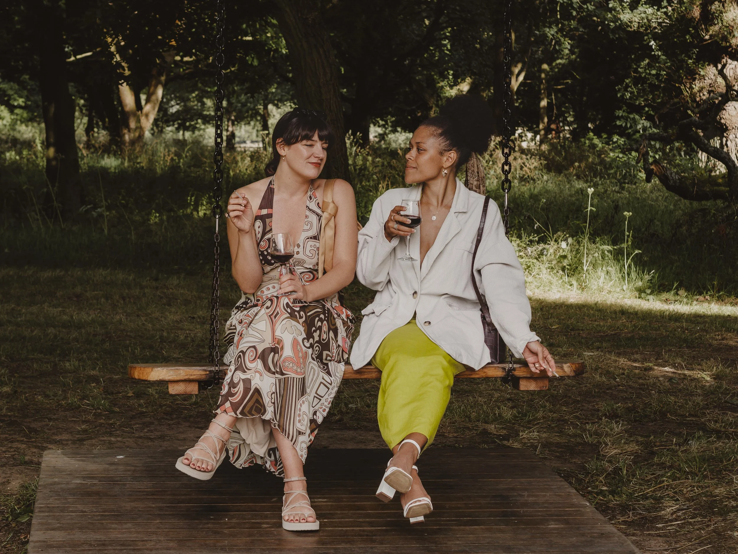 Two women sitting on a swing in a wooded park, holding glasses of red wine and engaging in conversation.