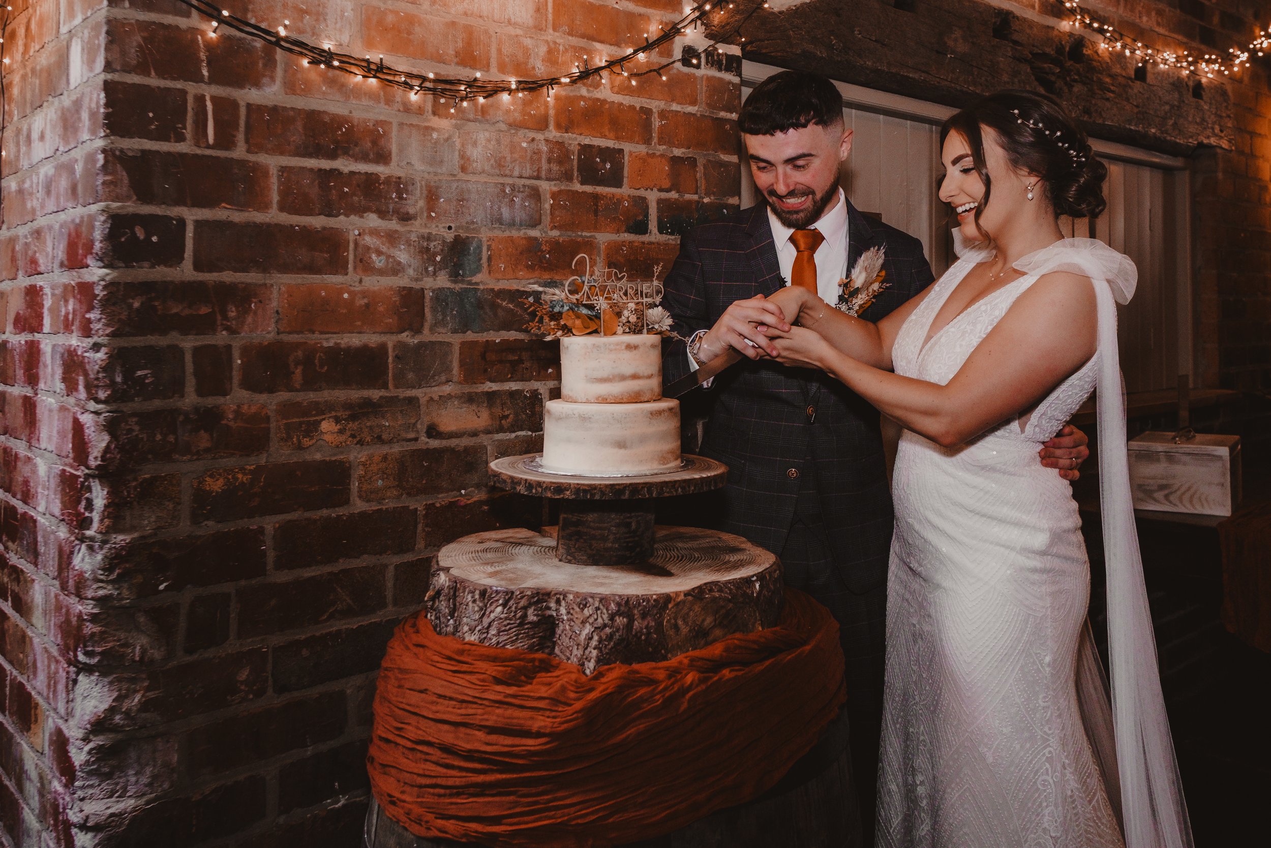 Bride and groom cutting a wedding cake in a rustic setting with exposed brick wall and string lights.