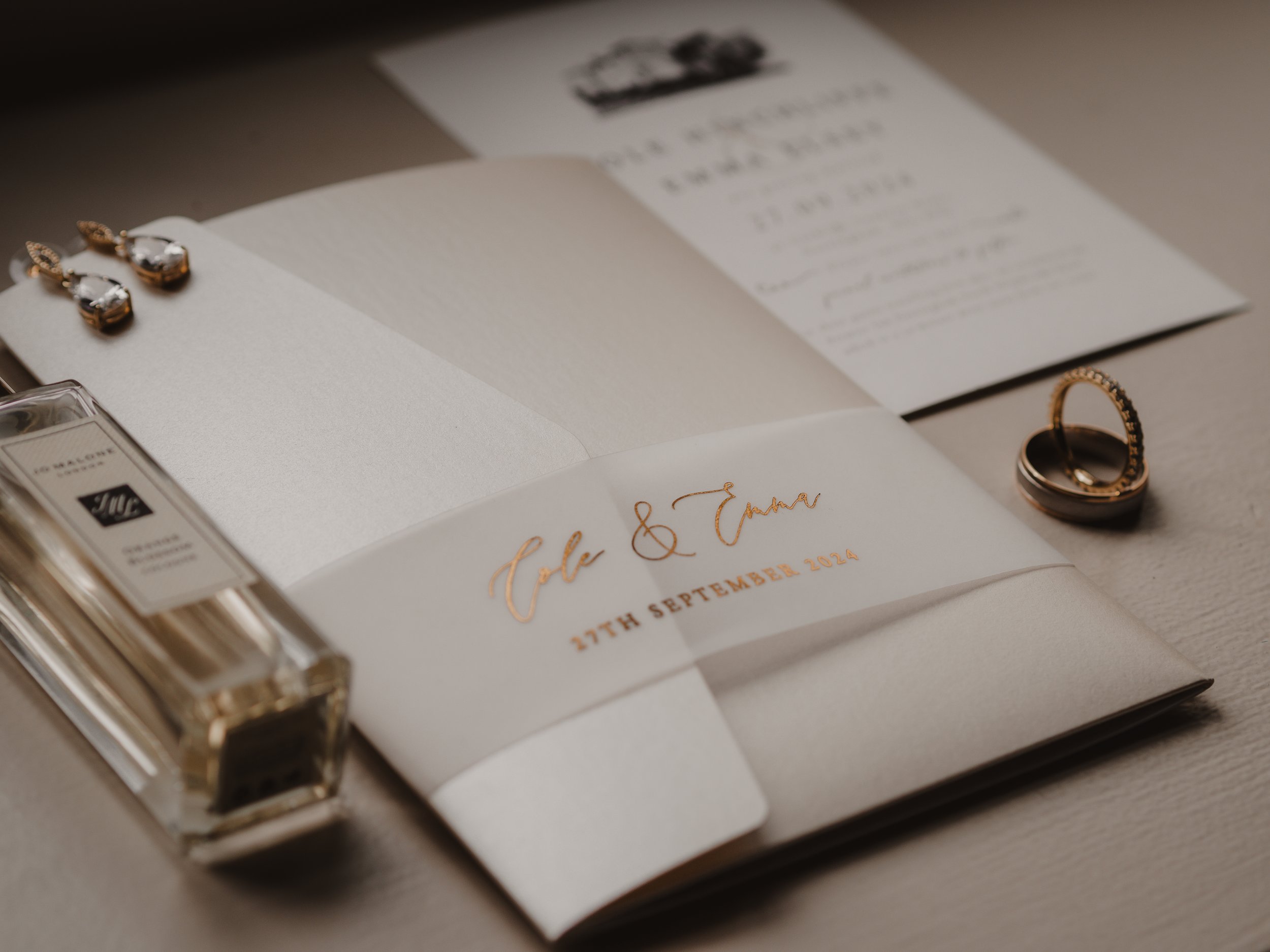 Wedding invitation with gold and white design, jewelry including earrings and rings, and perfume bottle.