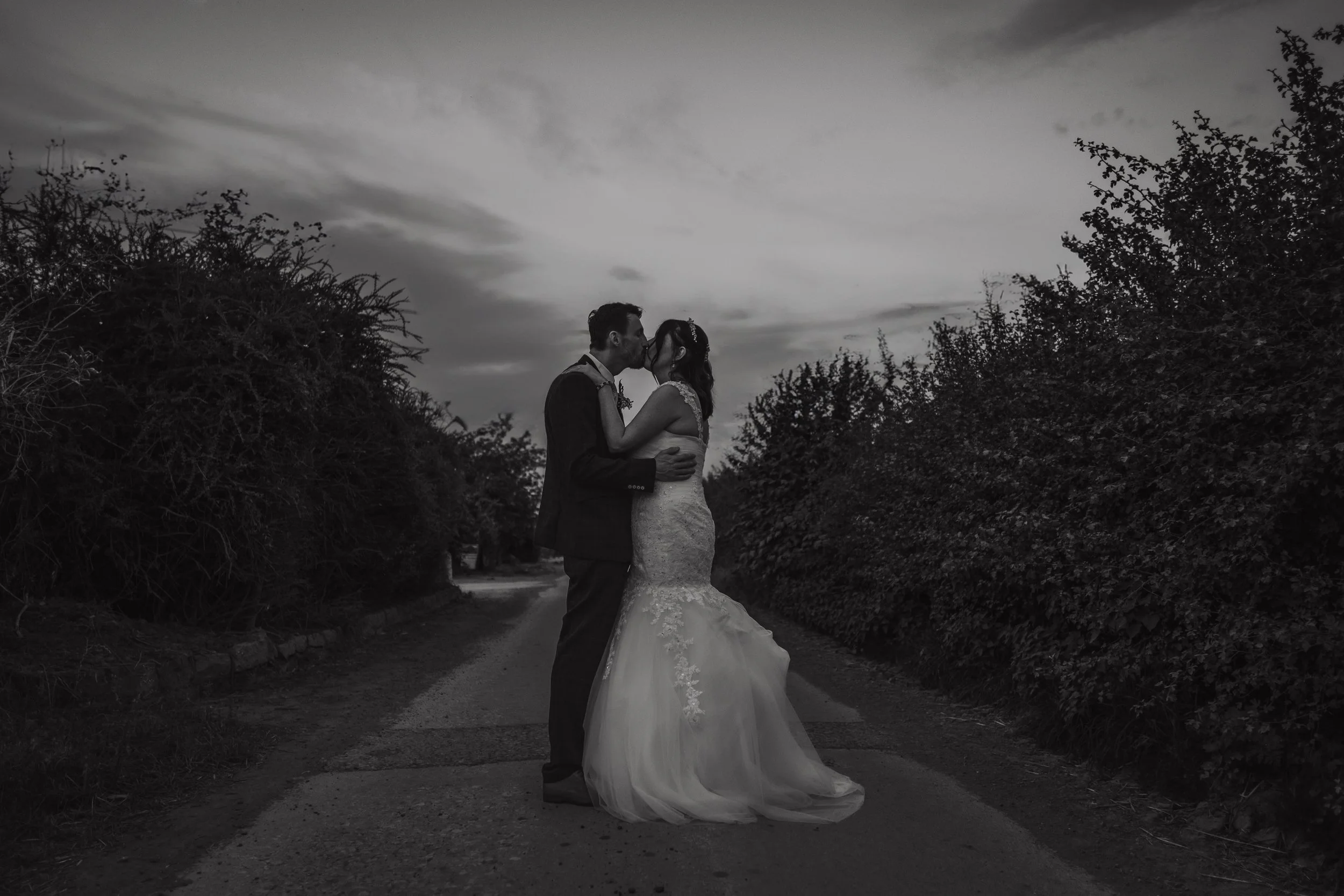 A couple in wedding attire sharing a kiss on a rural dirt road surrounded by bushes or small trees, captured in black and white during dusk or dawn.