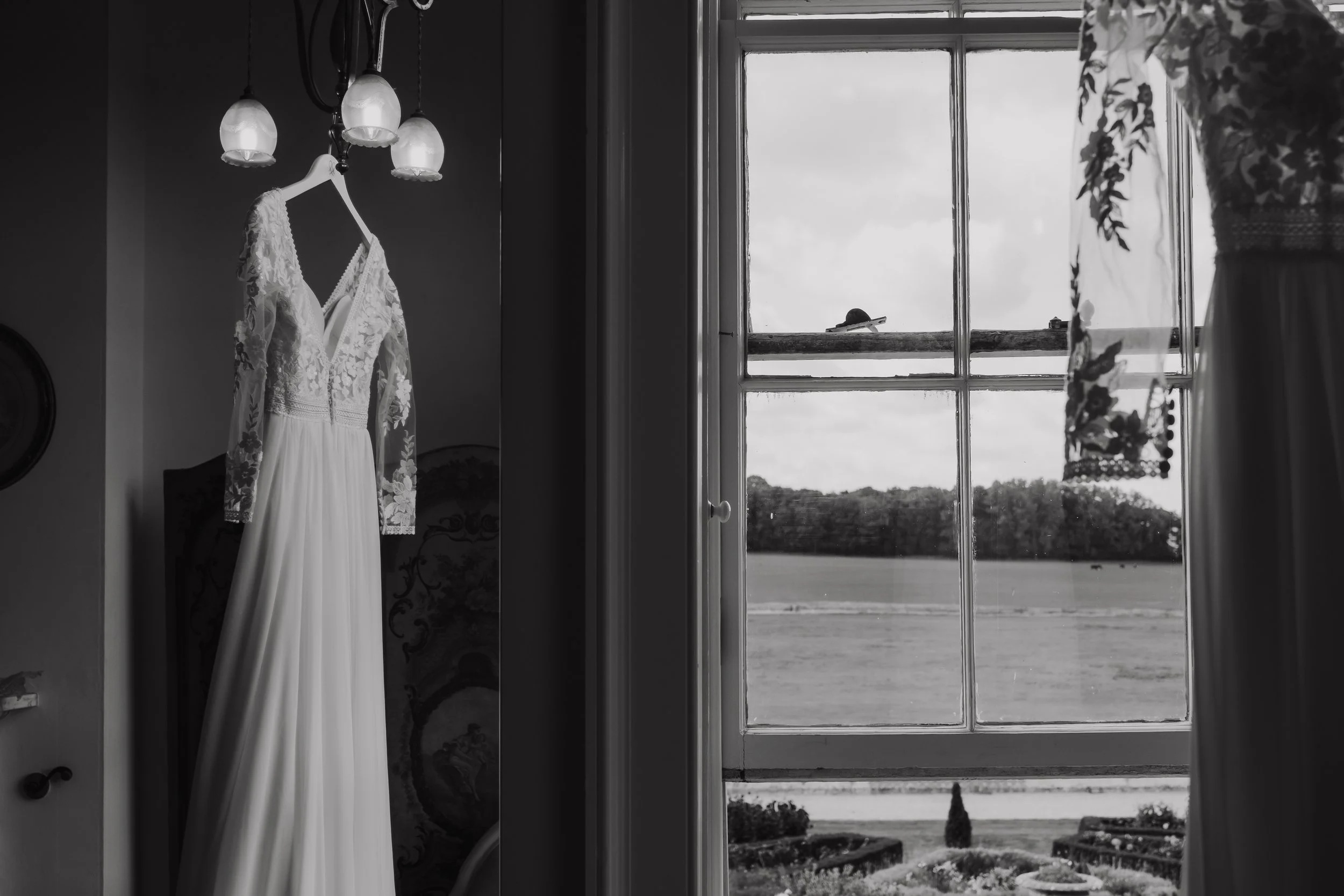 A wedding dress hangs on a hanger from a ceiling light fixture in a room, with a window showing an outdoor view of a field and trees.
