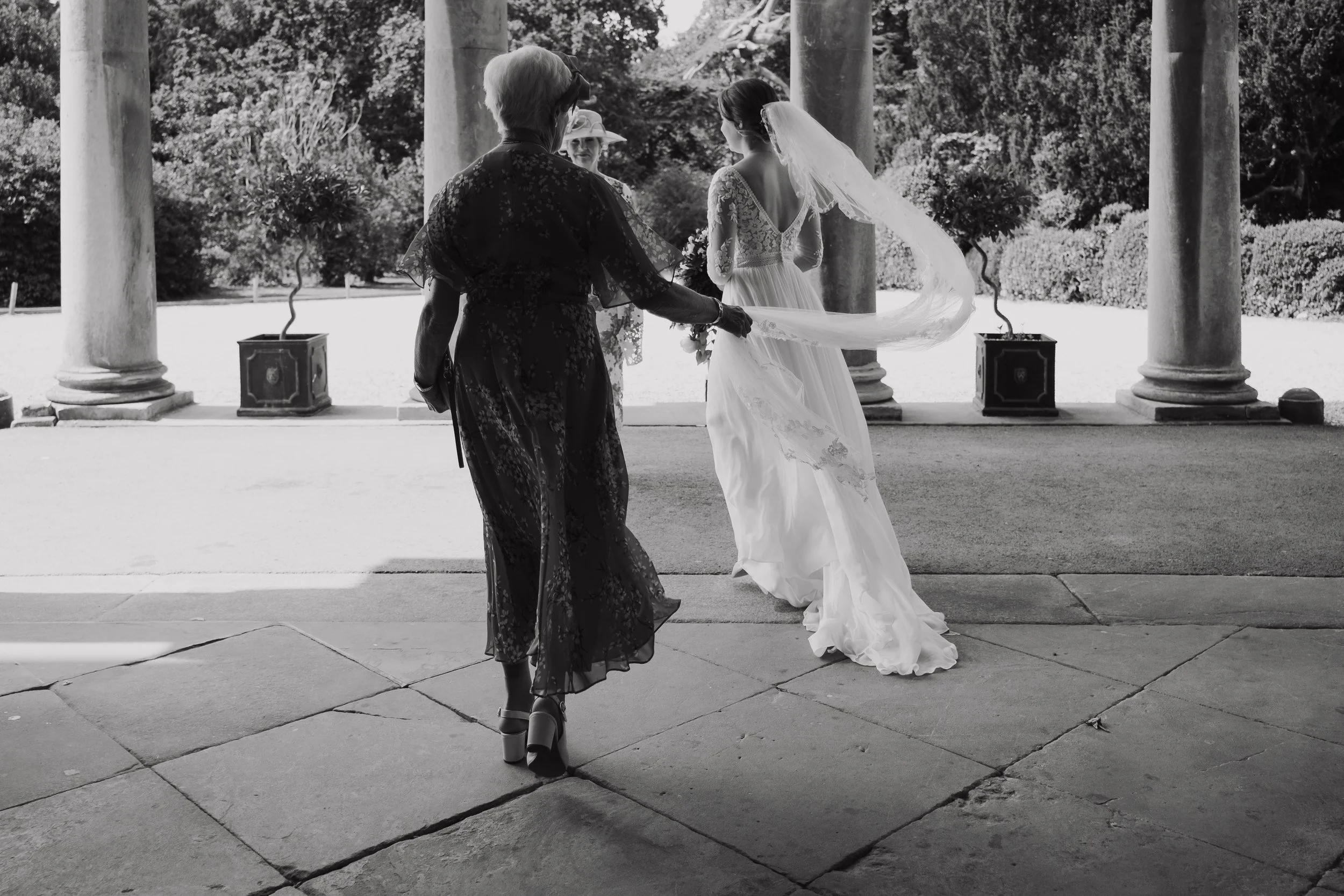 A bride in a wedding dress with a veil holding a bouquet, accompanied by her grandmother, outside under a pavilion with columns, trees, and bushes in the background.