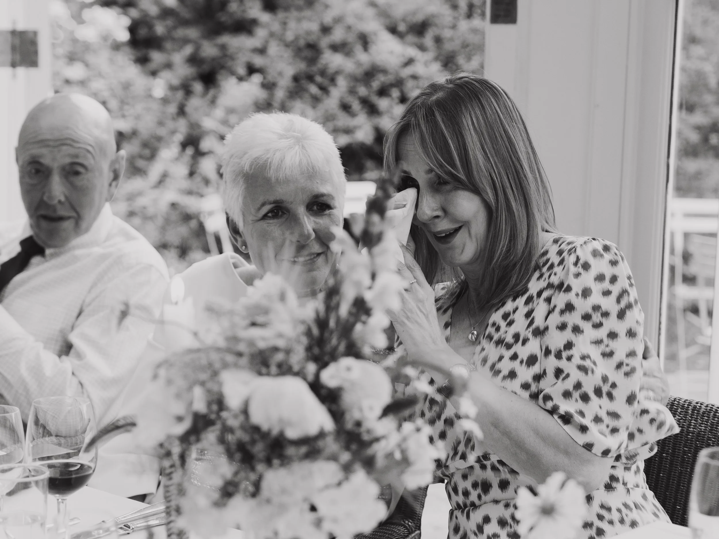 A black-and-white photo of three people sitting at a table with a flower arrangement. One woman is wiping away tears , while the other woman looks at her smiling and comforting her. A man is sitting on the left side of the image.