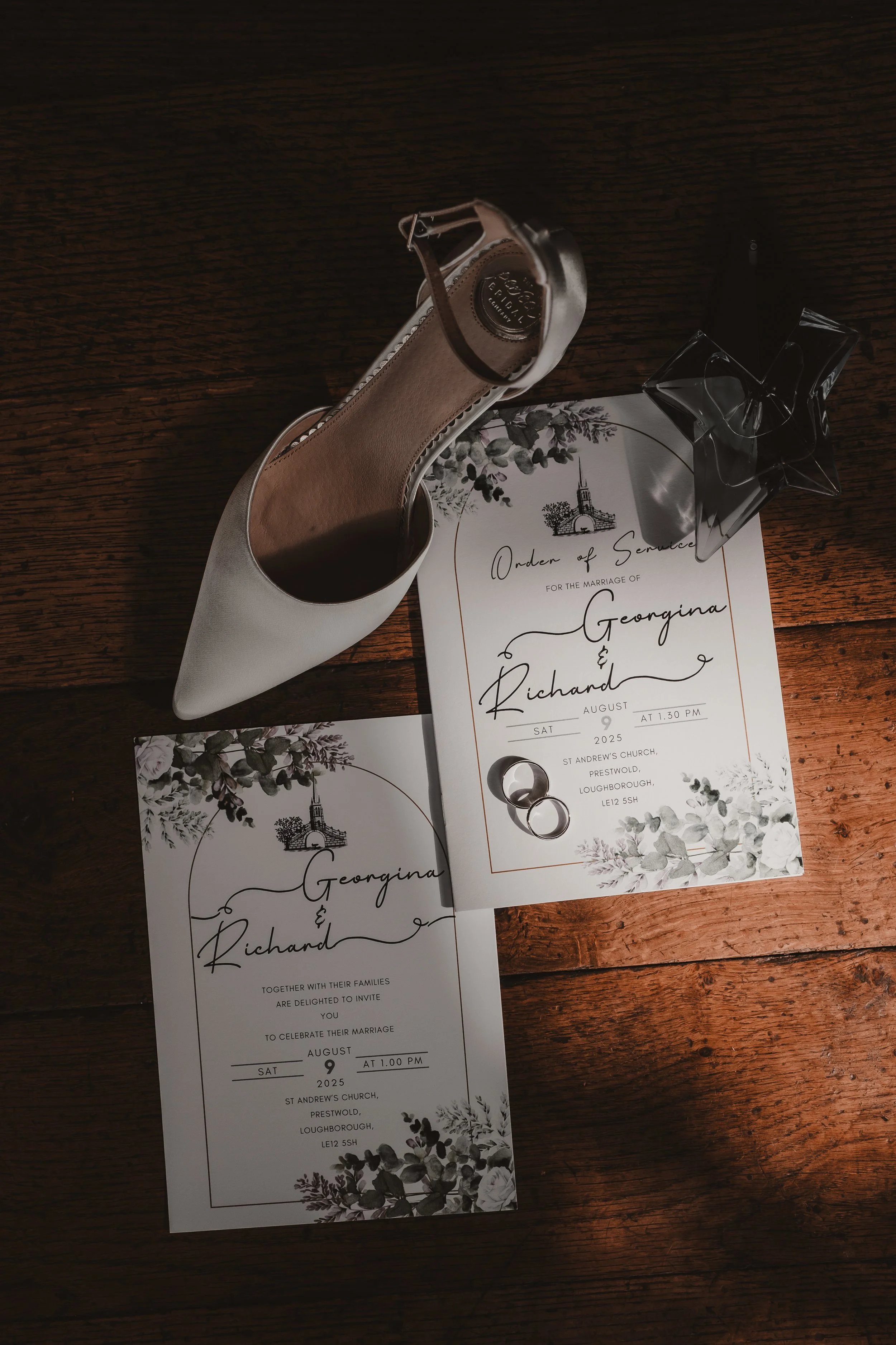 Wedding invitations with floral design, silver high heel shoe, wedding rings, and a star-shaped perfume bottle on a wooden surface.
