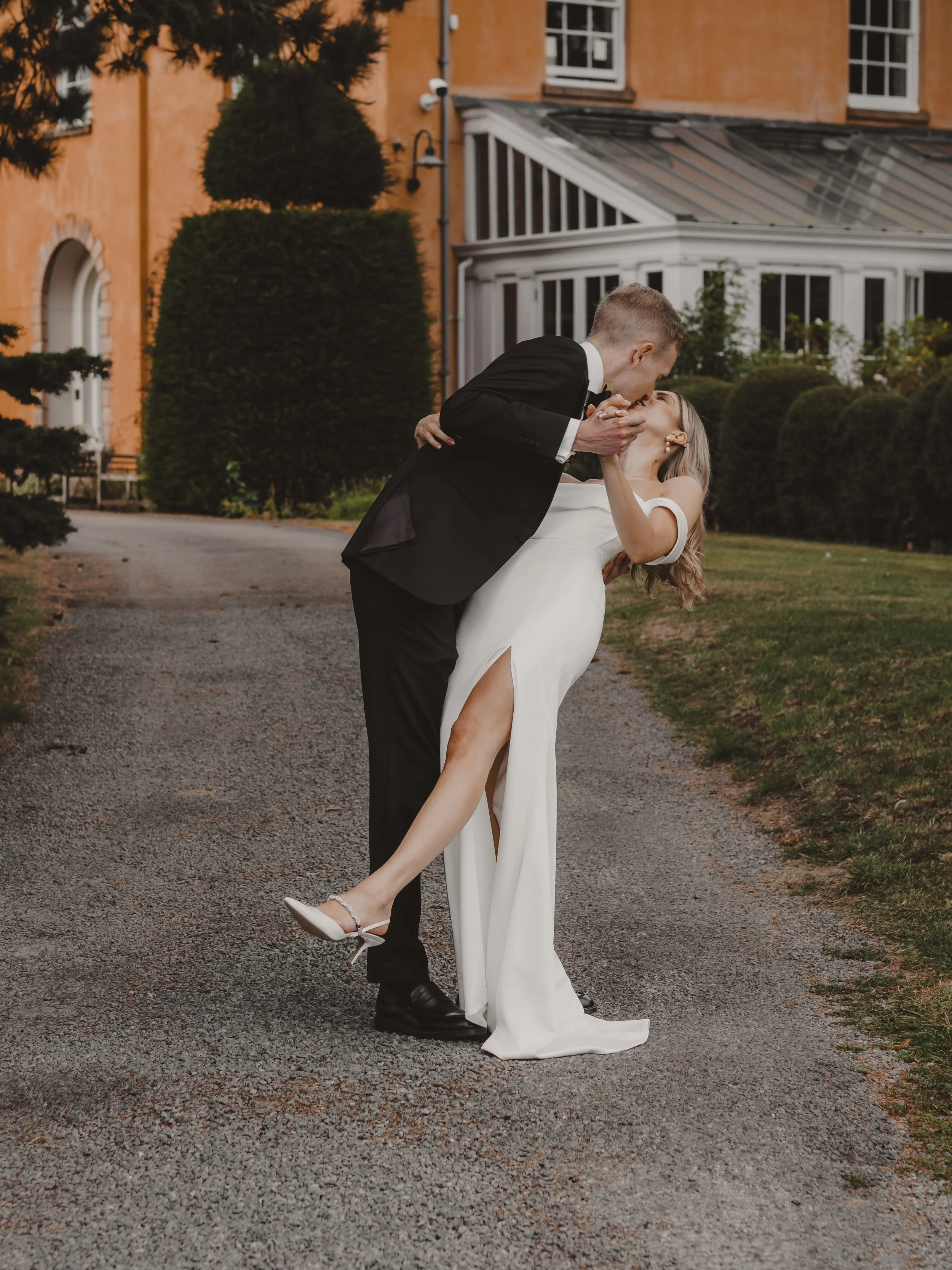 A couple in wedding attire sharing a kiss outdoors, with the man dipping the woman in front of Langar Hall