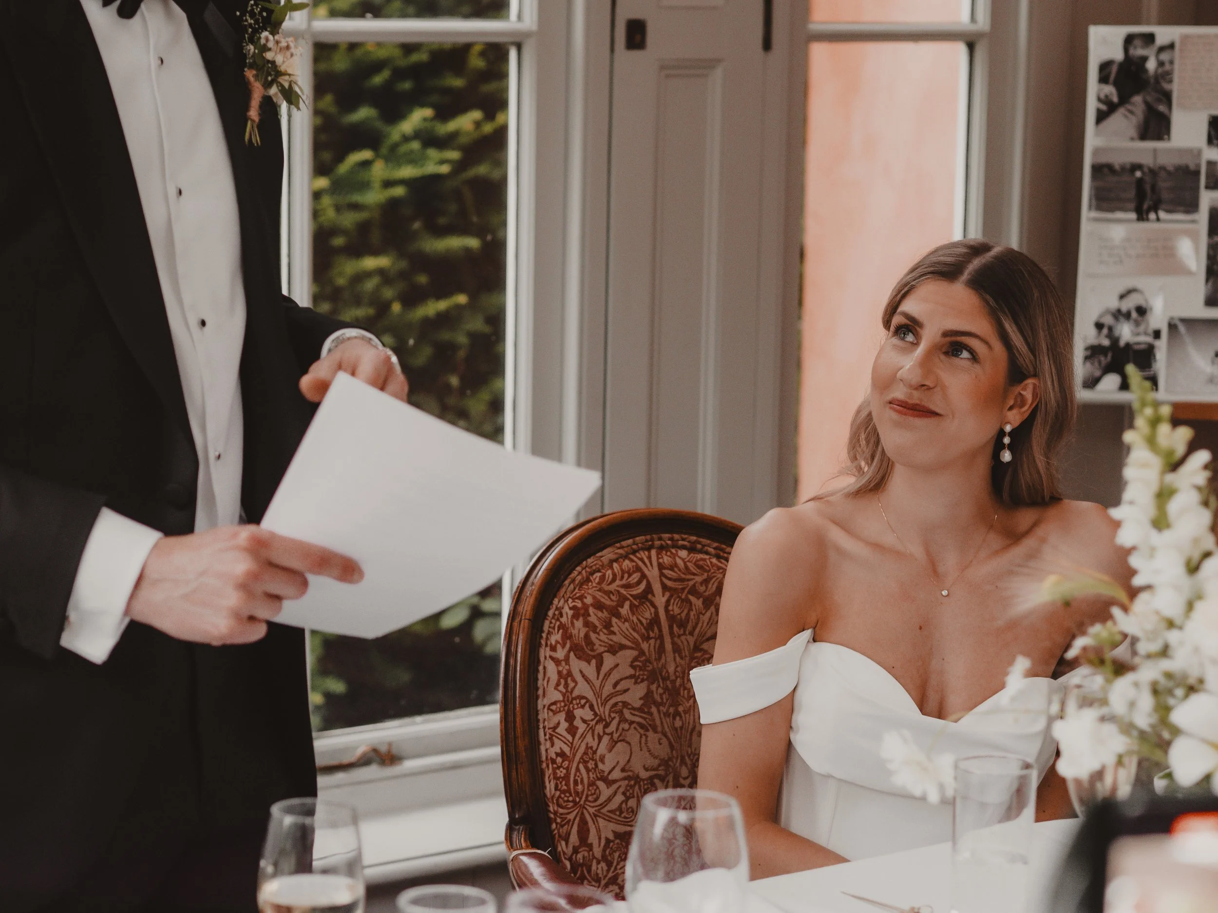 Bride is in an off-the-shoulder white dress sitting at a table during a wedding reception, looking up at the groom in a black tuxedo who is holding his speech.