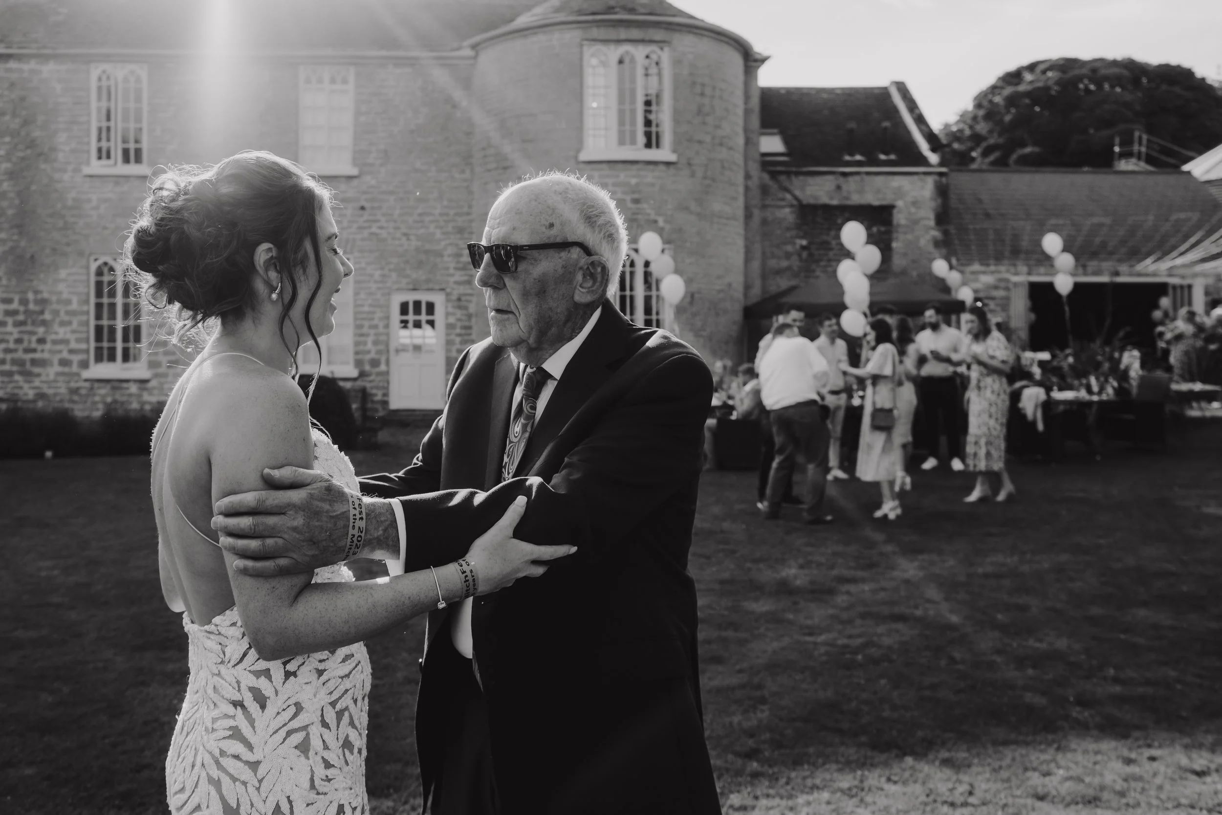 A young woman in a lace dress is holding hands and smiling at an elderly man wearing sunglasses and a suit, both are outside near a brick building at a celebration with guests and balloons in the background.