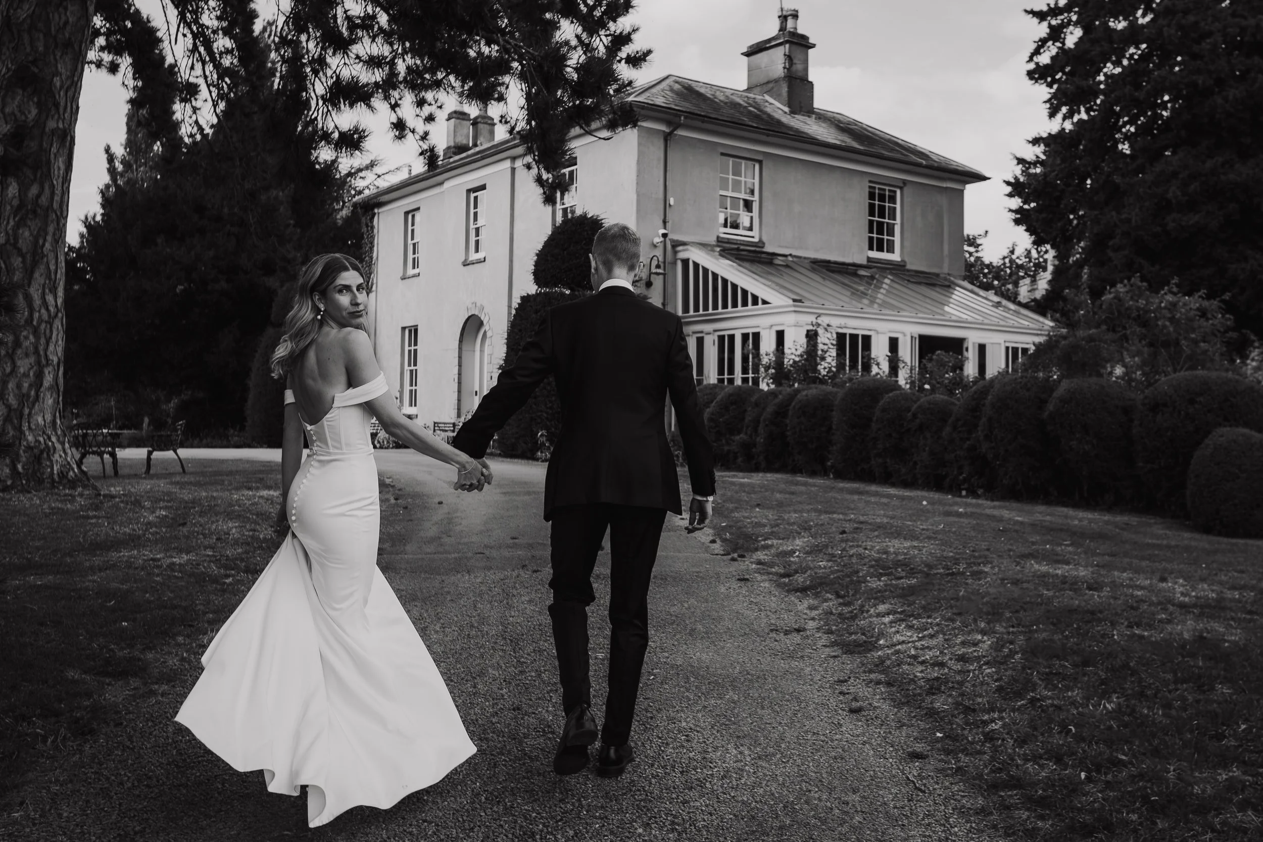 A black and white photo of a bride and groom walking hand in hand on a gravel path in front of a large house with a conservatory and manicured bushes. The bride is wearing a strapless, form-fitting wedding gown and looking back over her shoulder, whi