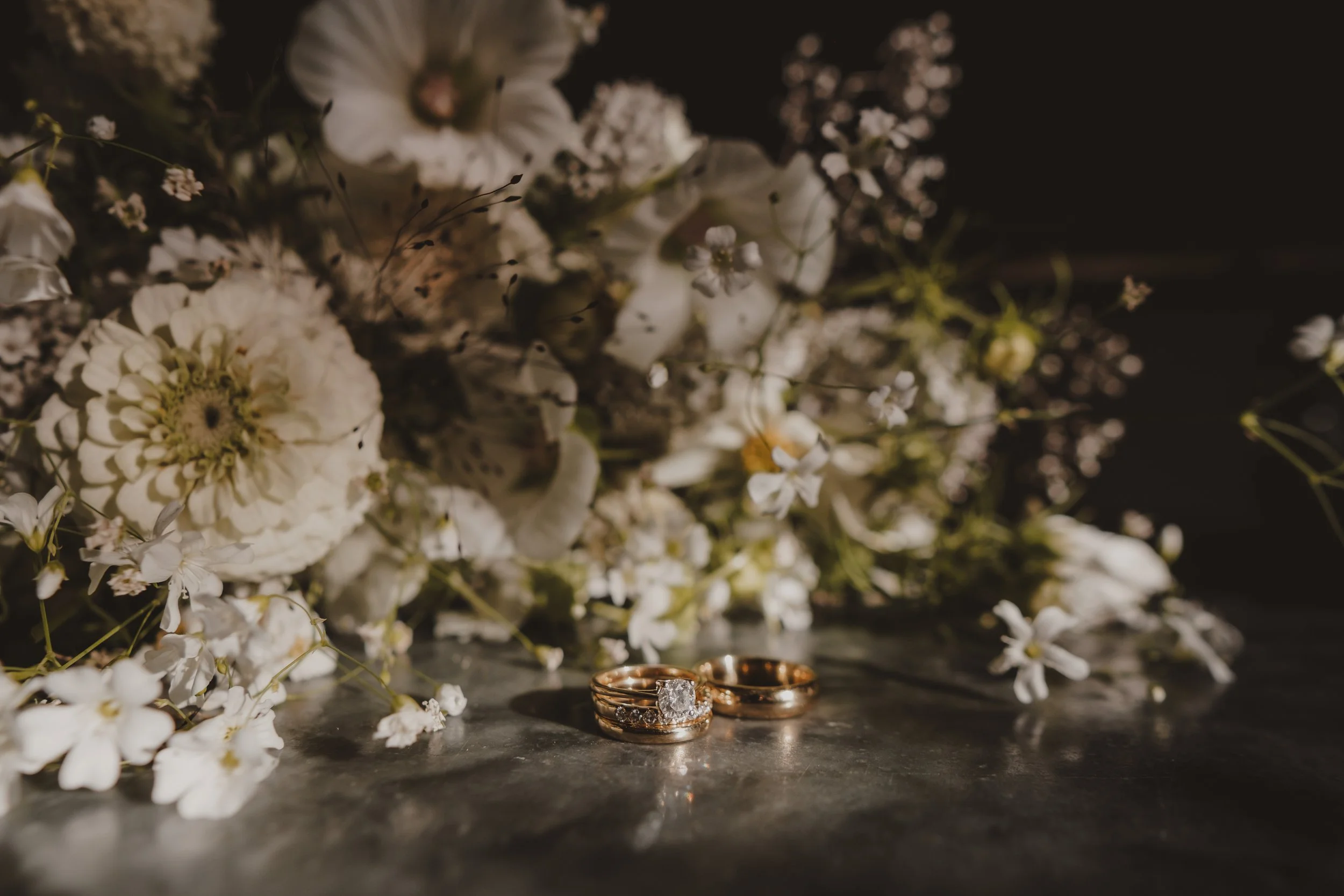 Close-up of wedding or engagement rings surrounded by white flowers and dark background.