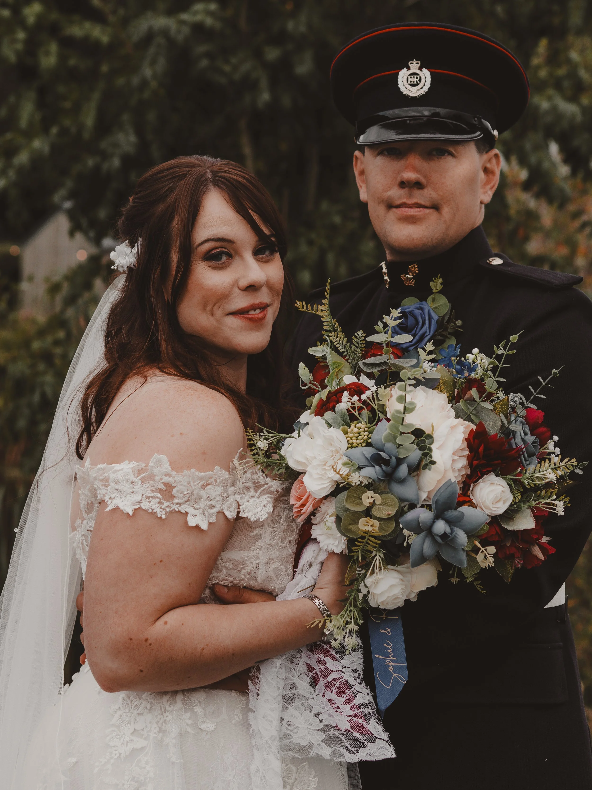A bride and groom in wedding attire, holding a bouquet of white, red, and blue flowers with greenery, outdoors with blurred trees in the background.