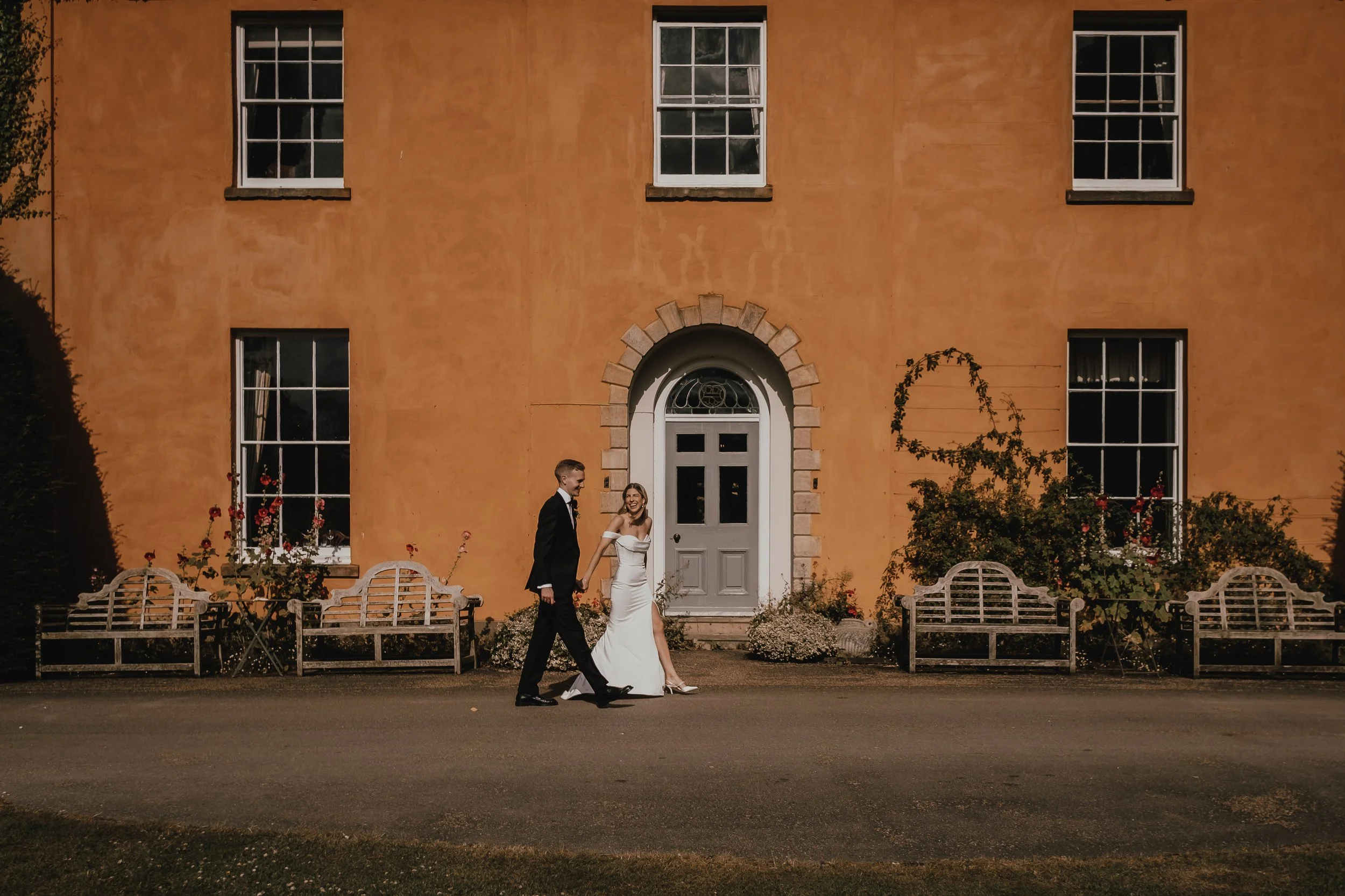 A newlywed couple walking hand in hand in front of a large orange building with white window frames and a gray door. The bride is in a white wedding dress and the groom in a black tuxedo.