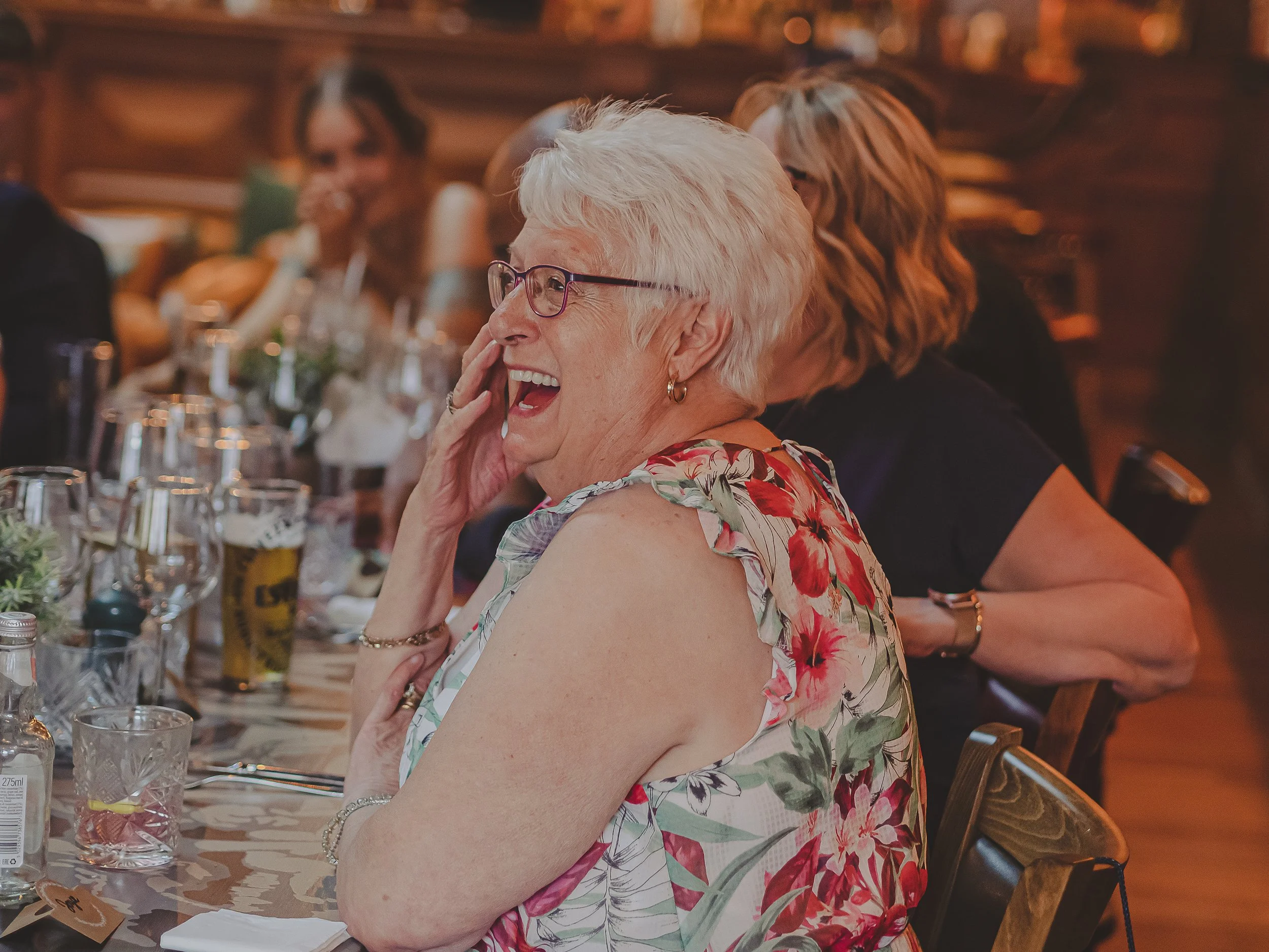 An elderly woman with short white hair and glasses, wearing a sleeveless floral dress, is laughing and touching her face during a speeches at a wedding at Cosy Club Nottingham .
