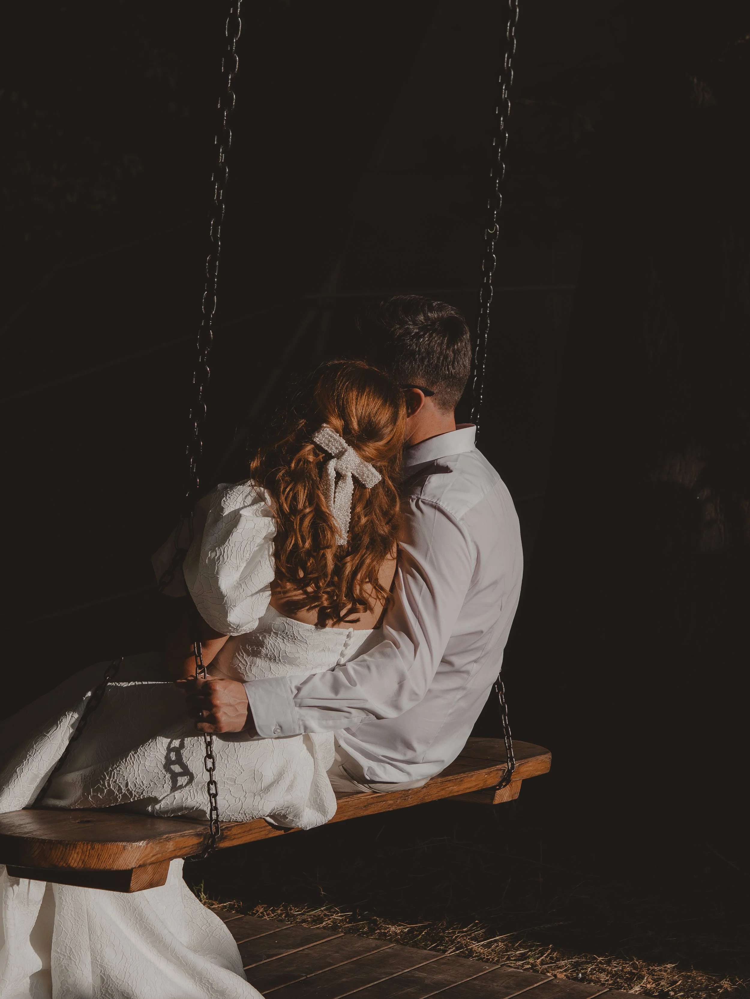 Wedding couple sitting on a wooden swing, embracing each other at night, with dark background and faintly visible trees.