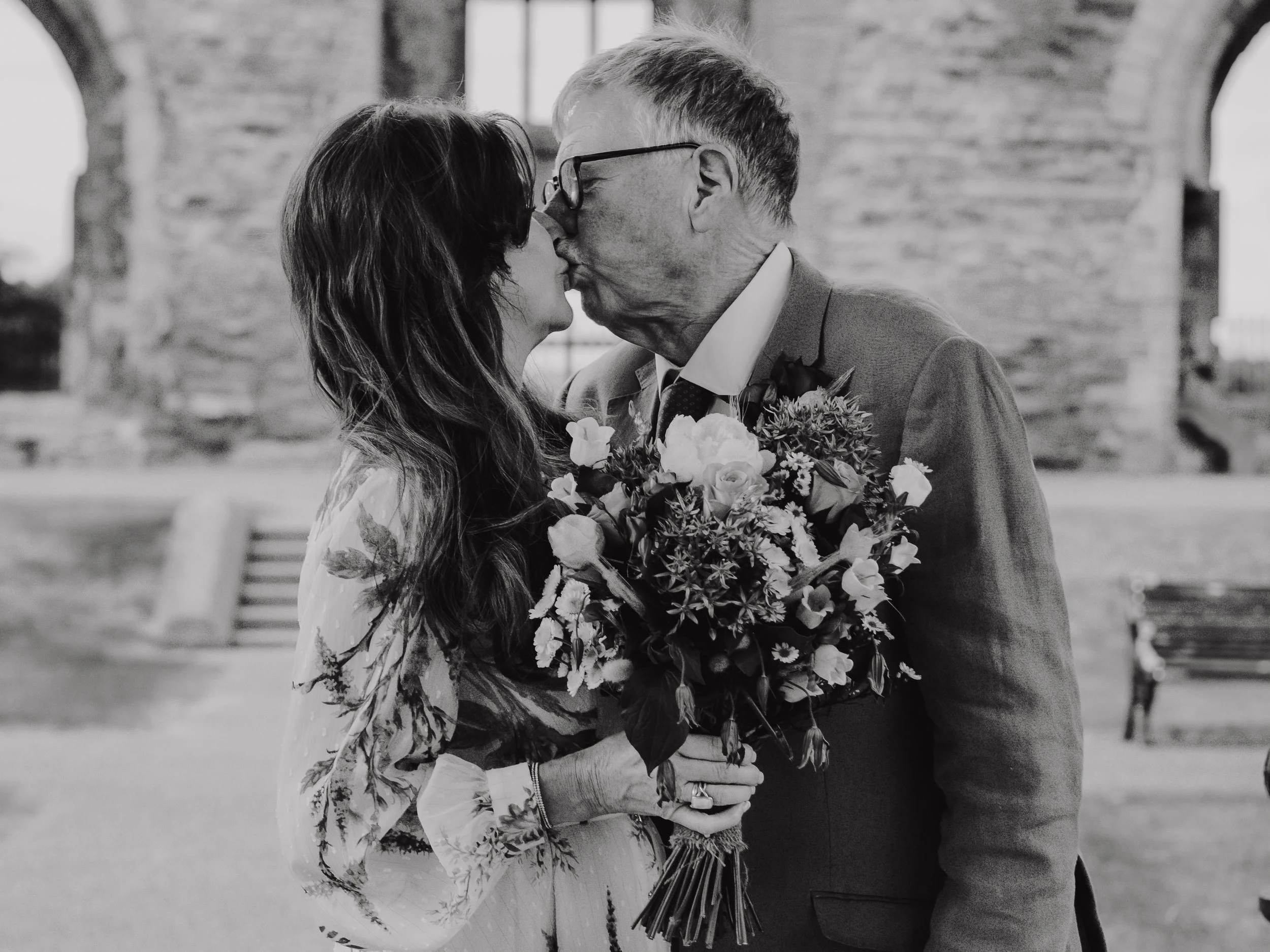 Bride and groom are sharing a kiss outdoors with the woman holding a bouquet of flowers, and stone arches in the background.