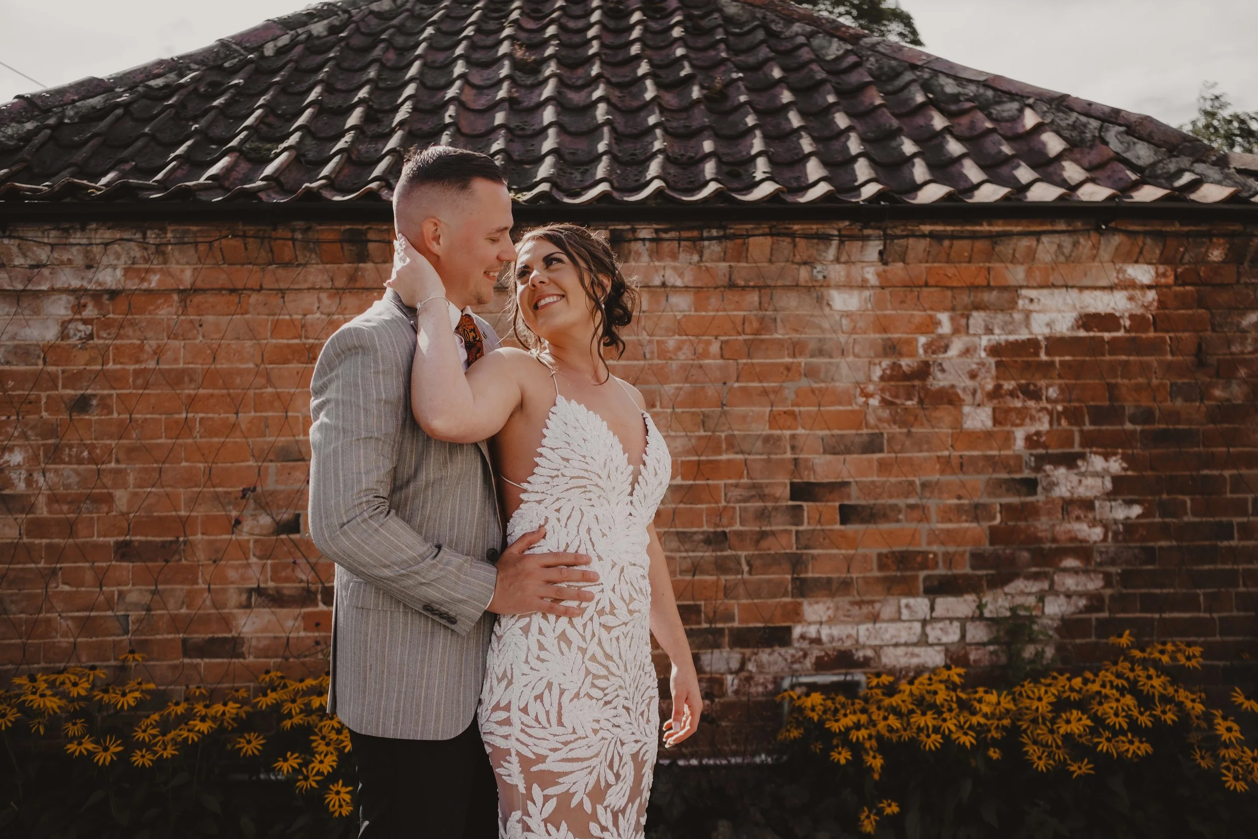 A couple on their wedding day standing closely together, smiling and looking at each other, in front of a brick wall with yellow flowers at the base.