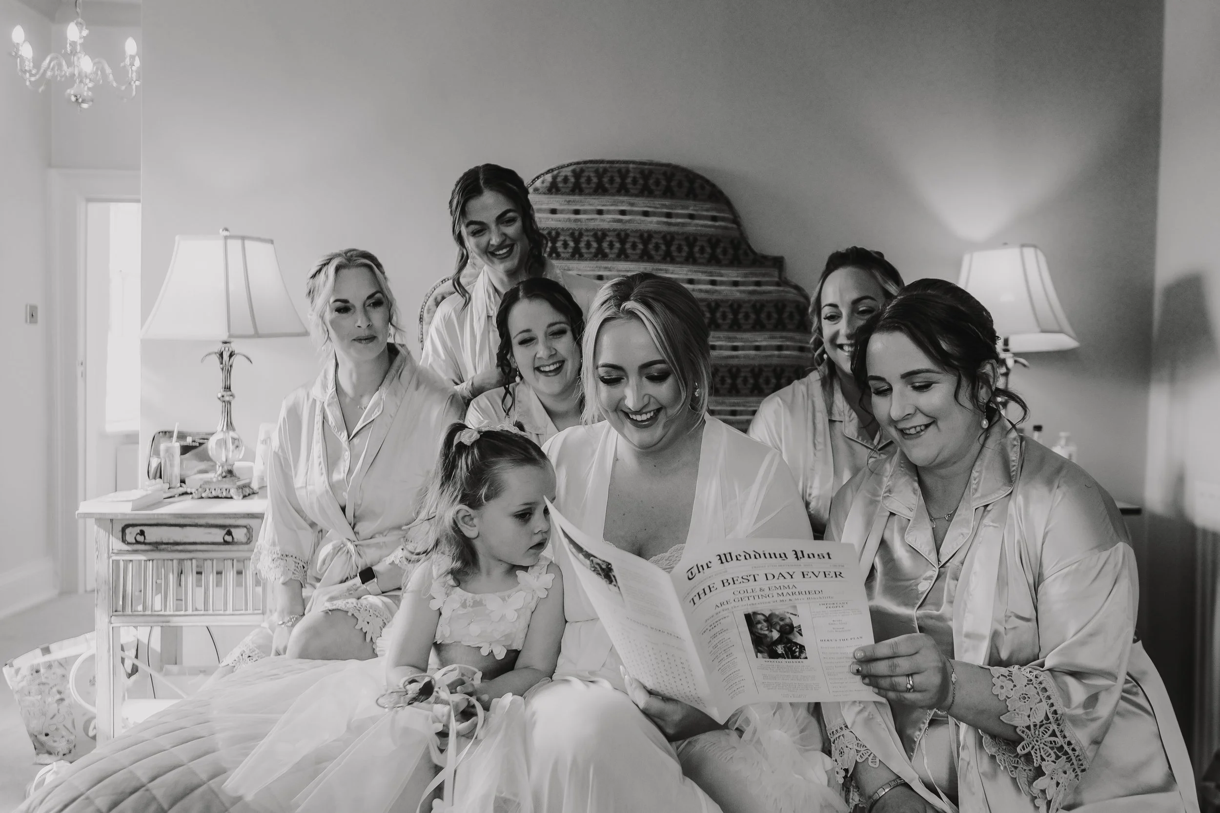 Group of women and a young girl in bridal robes sitting on a bed, looking at a newspaper titled 'The Wedding Post' with a headline 'THE BEST DAY EVER'. They are smiling and appear happy,  getting ready for a wedding.