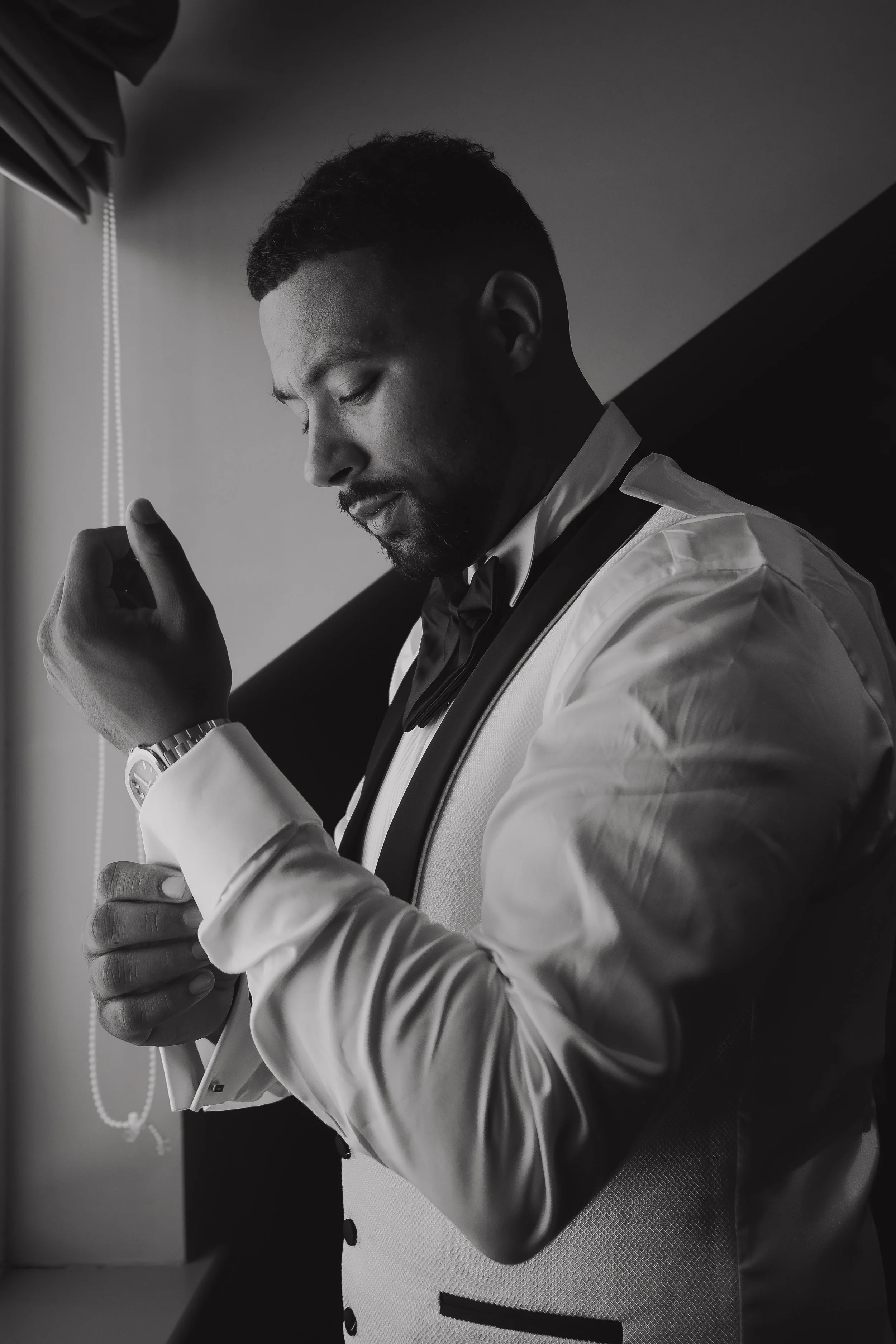 A man adjusting his cufflinks while dressed in a tuxedo with a bow tie, standing near a window with natural light illuminating his face.