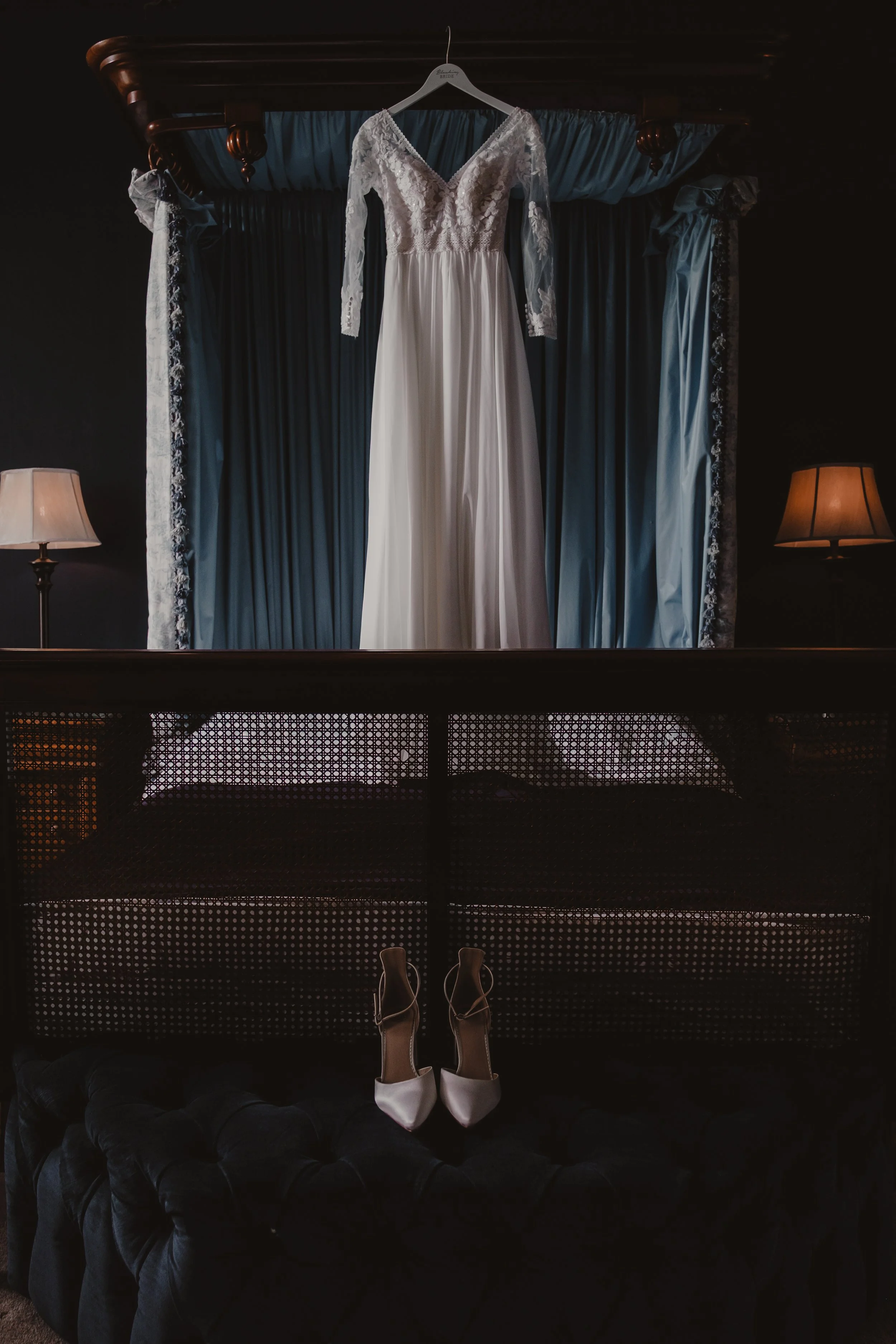 Bridal gown hanging from a wooden canopy with blue curtains, two lamps on bedside tables, and a pair of white high-heeled shoes on a black ottoman below.