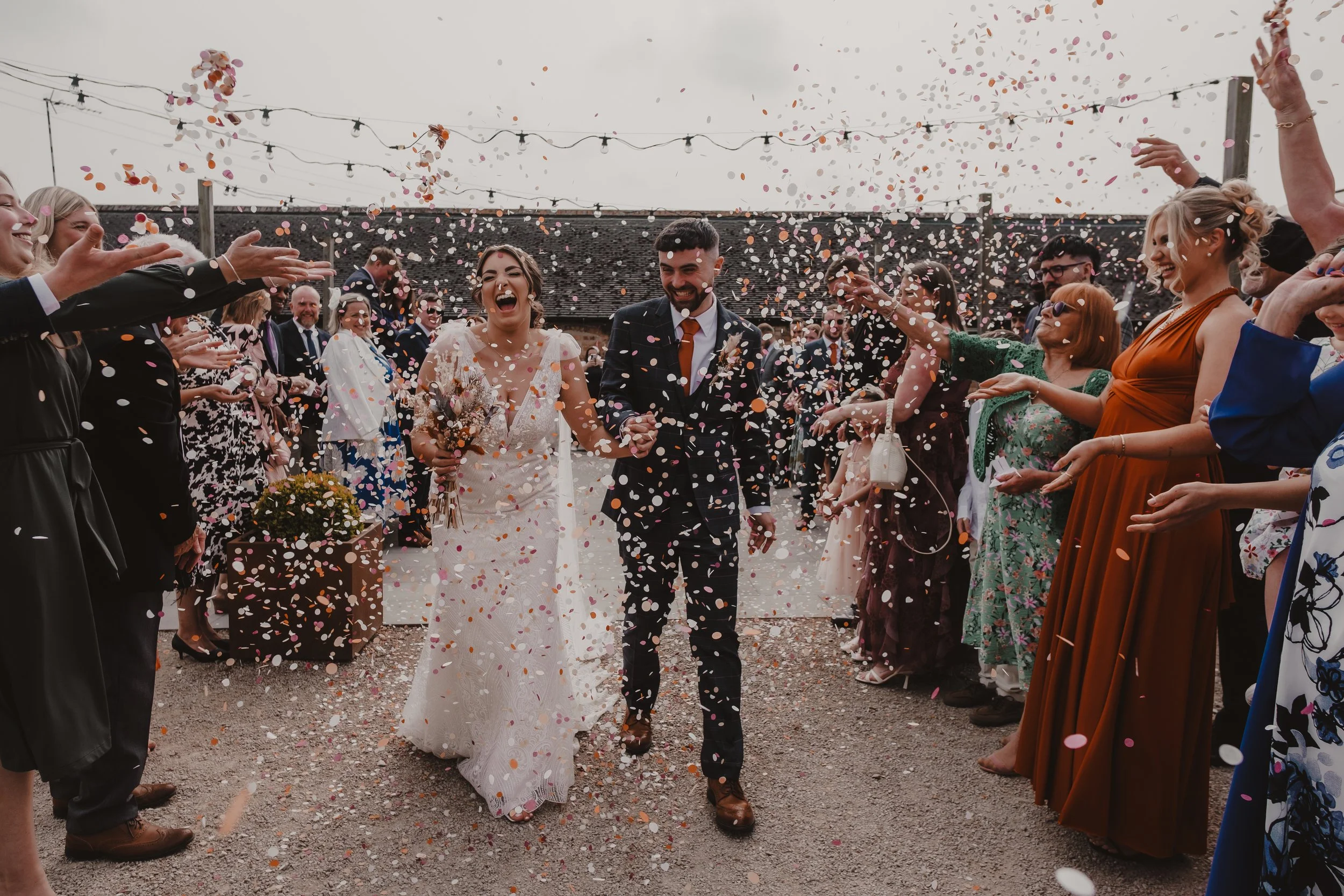 Bride and groom walking hand in hand through guests throwing confetti at an outdoor wedding celebration.
