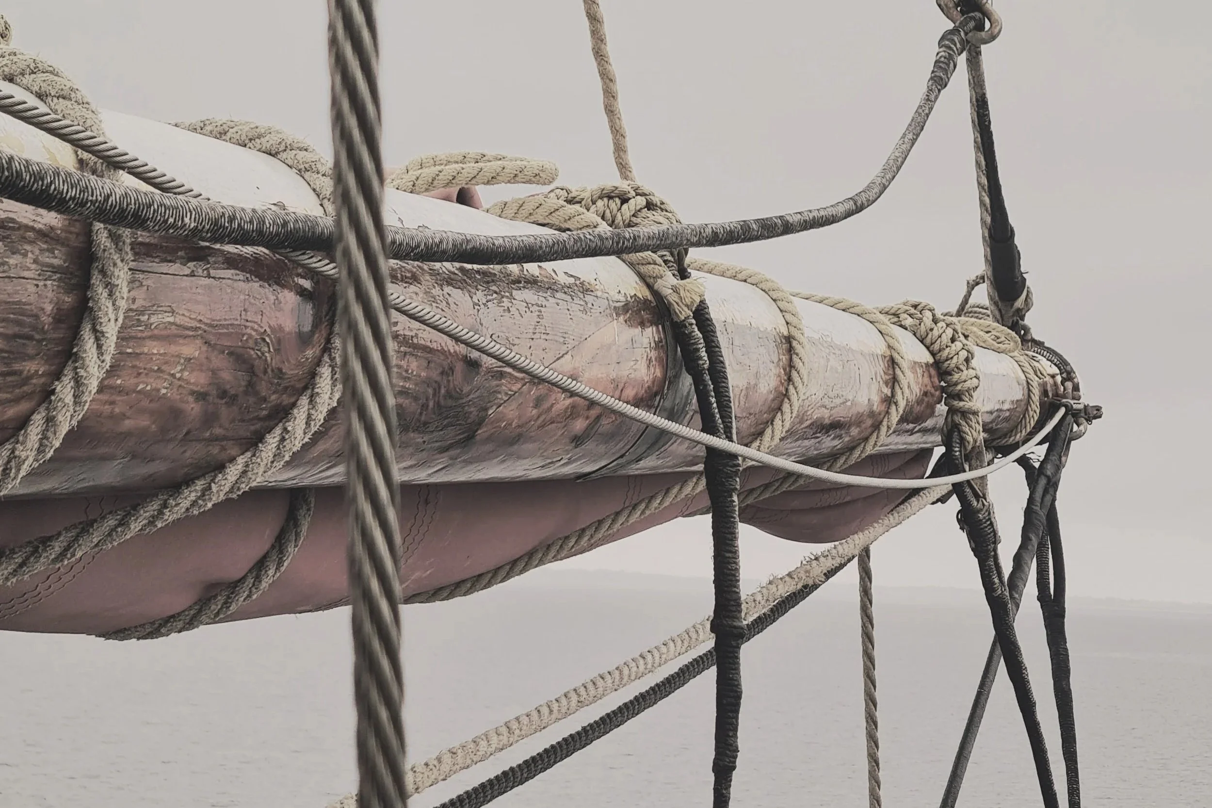 Close-up of the mast and rigging of a boat or ship at sea, with water in the background and a cloudy sky.