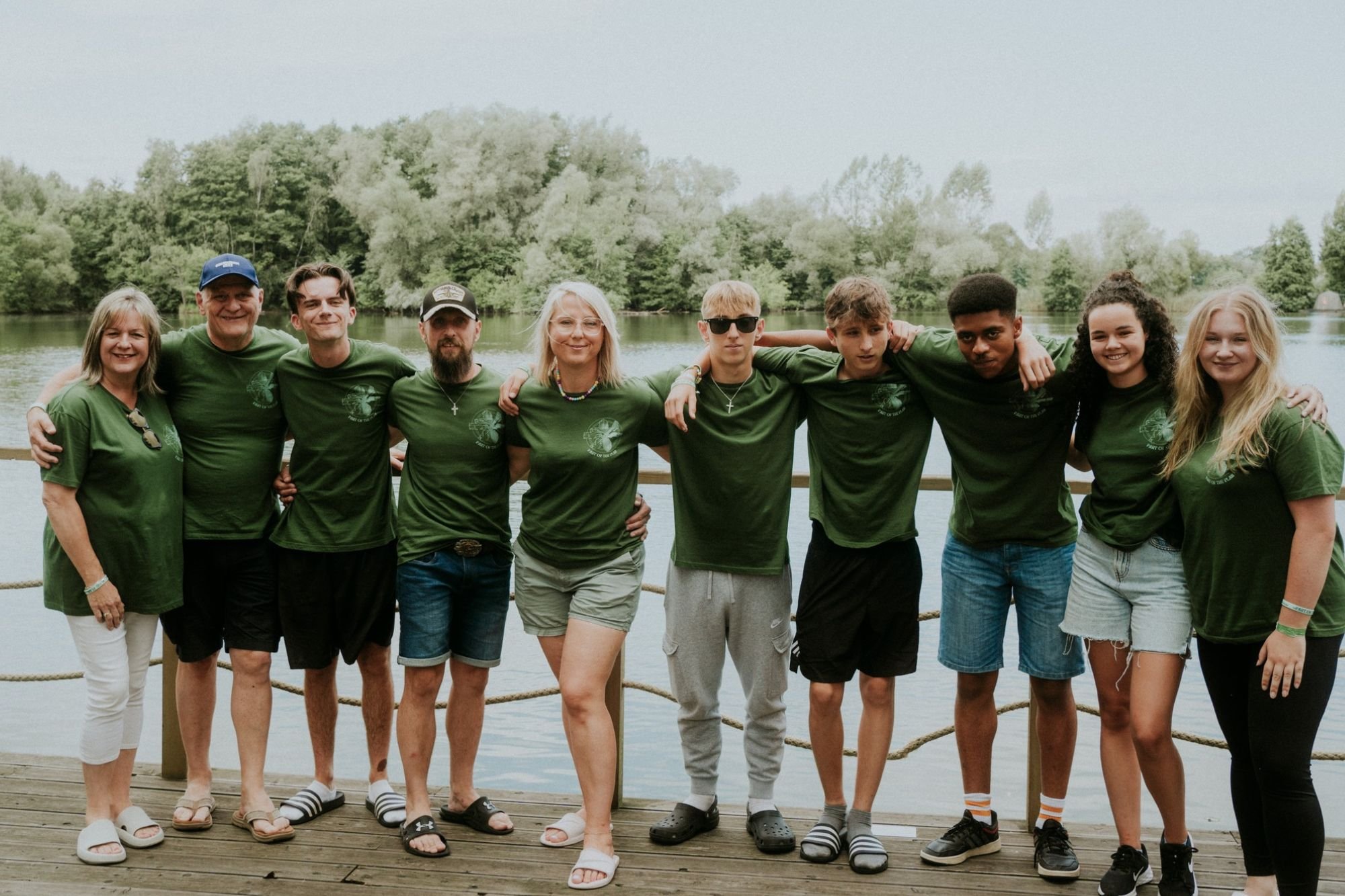 Group of ten people standing on a wooden dock by a lake, with trees in the background, all wearing green shirts.