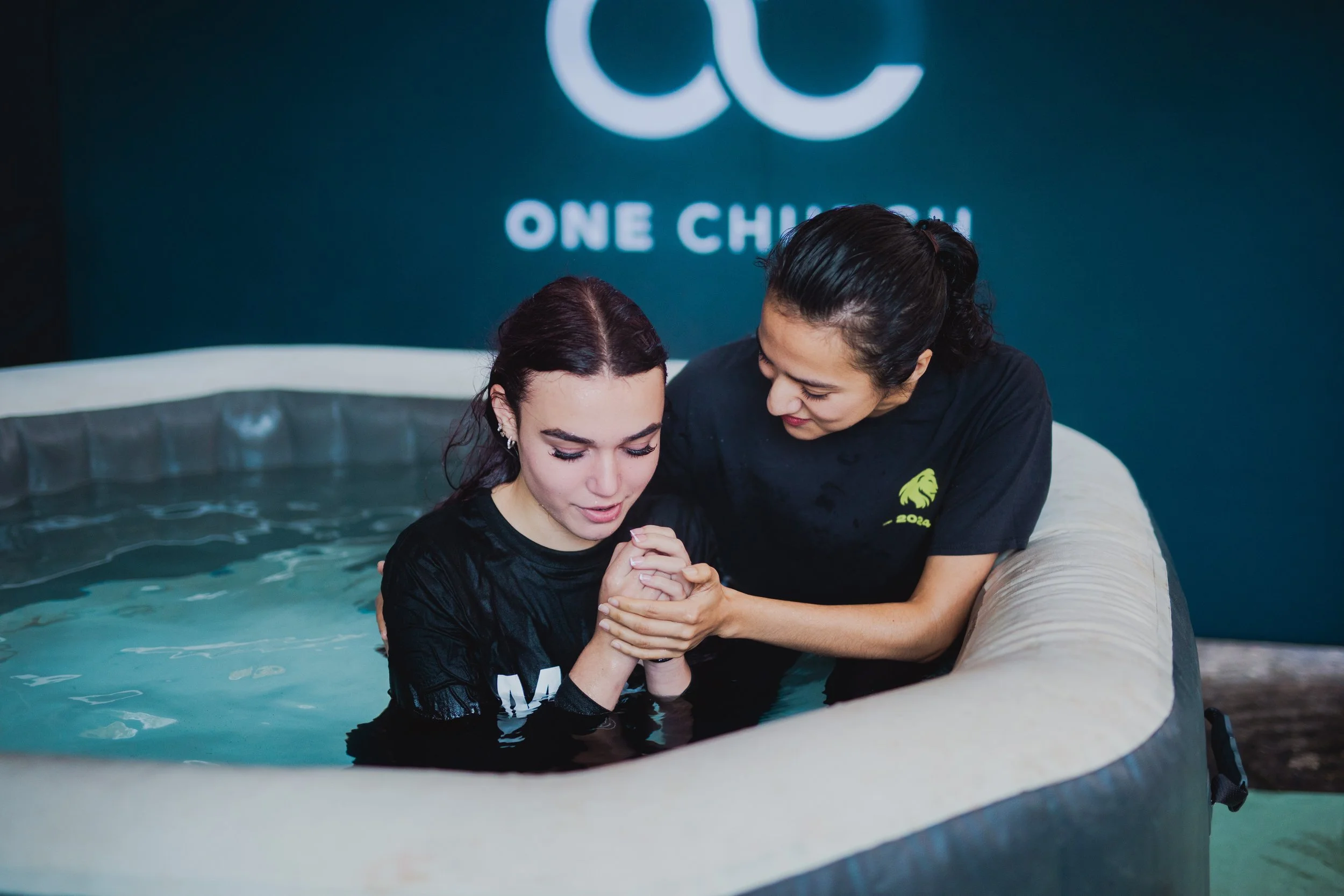 Two women in black shirts involved in a baptism ceremony, with one in the water and the other assisting her, in front of a dark blue wall with a large white logo and the words 'One Church'.