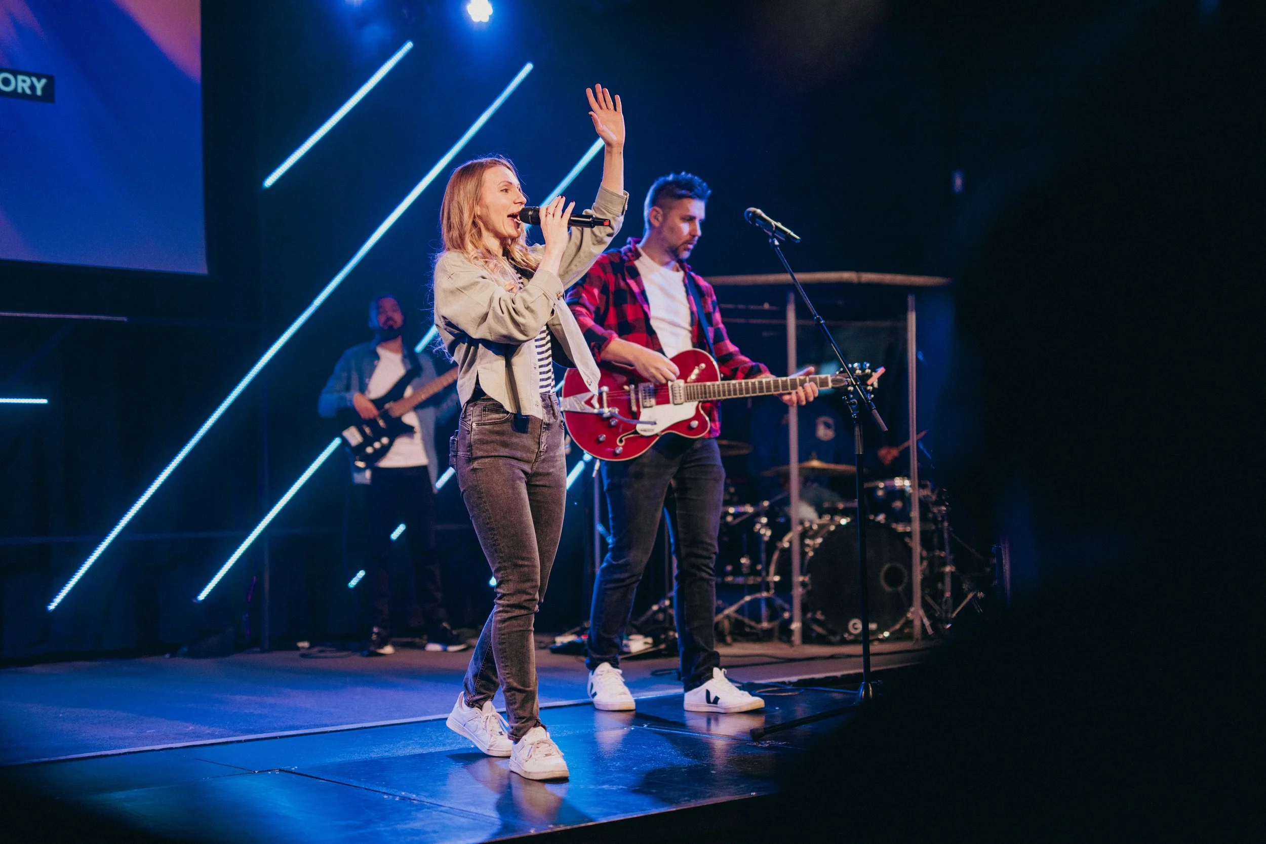 A band performs on stage with a woman singing into a microphone, accompanied by a male guitarist, with other band members in the background.