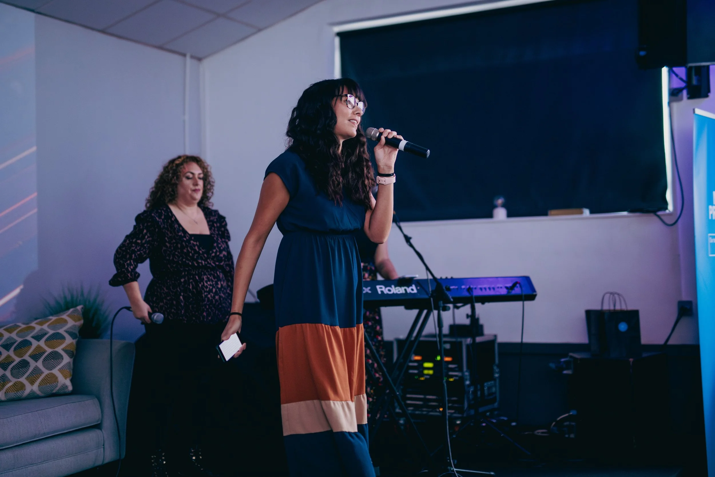 A woman with glasses singing into a microphone on a stage with a keyboard behind her, and another woman holding a mic and a phone nearby, in a room with a gray couch and decorative pillow.