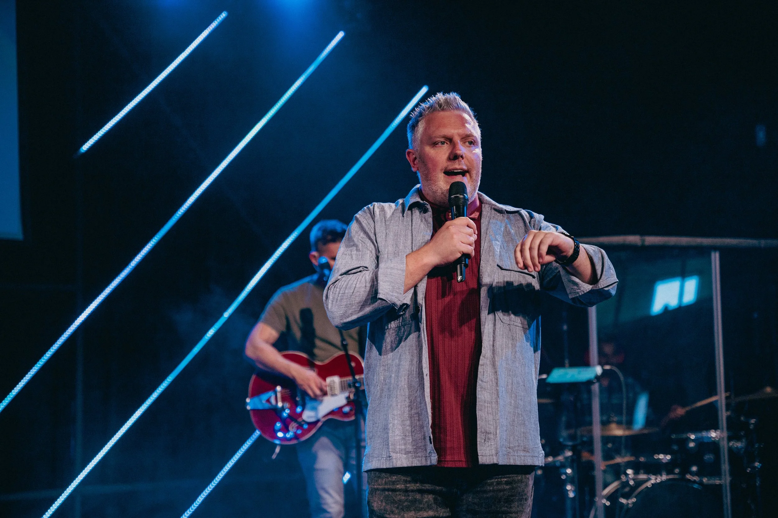 A man singing into a microphone on stage, with a guitarist playing in the background, neon lights overhead, and a dark background.