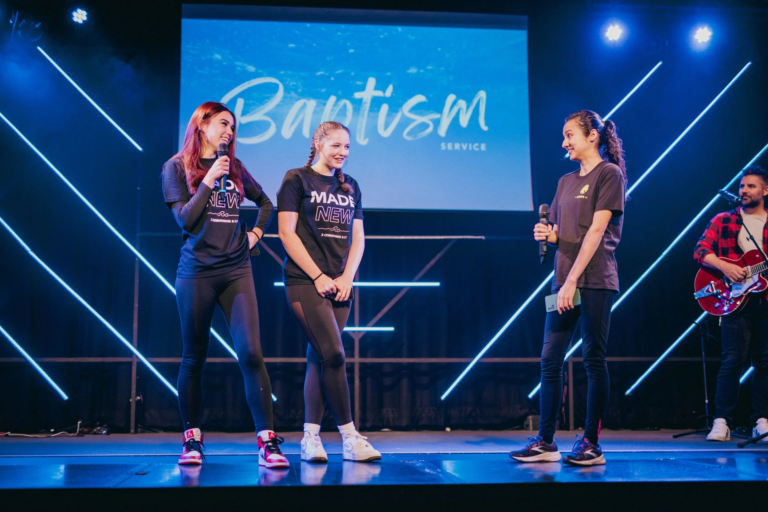 Four young women on stage during a baptism service, with three holding microphones and one with a guitar, in front of a large screen displaying 'Baptism Service' and blue neon lights.