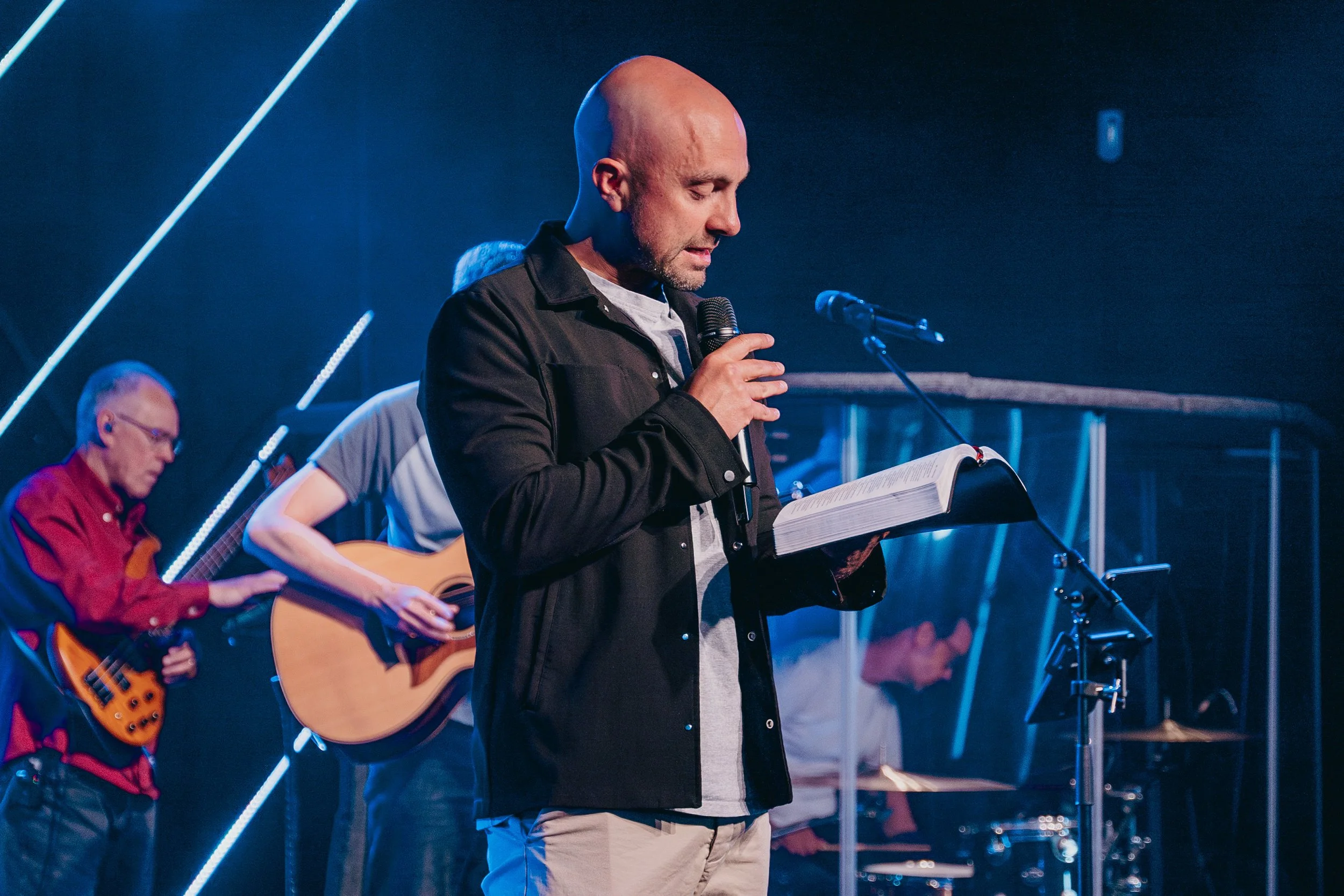 A man with a shaved head and beard reads from a book while holding a microphone on stage, with two other men in the background playing guitars and drums.