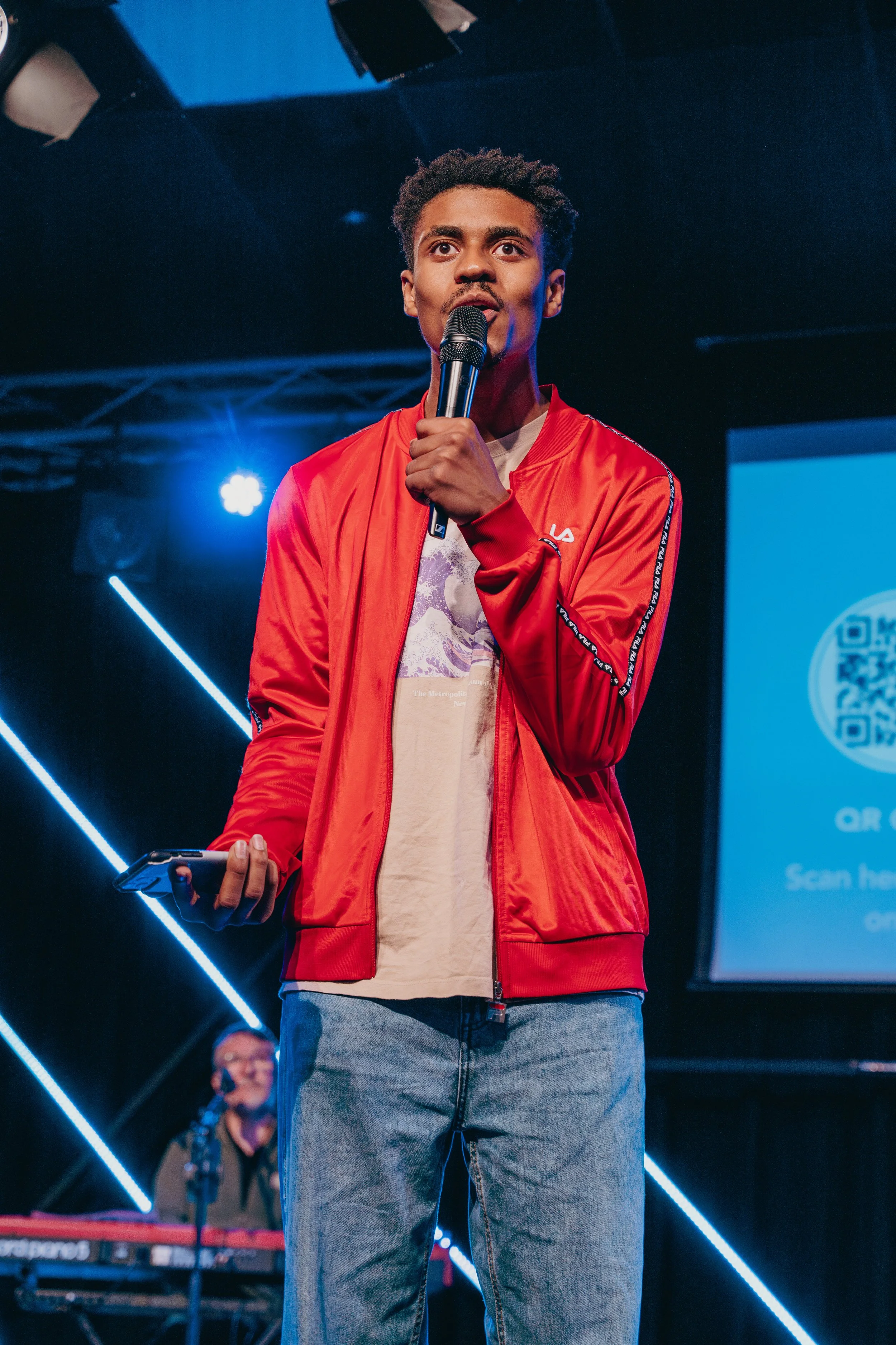 A young man in a red jacket speaking into a microphone on stage with another person playing a keyboard in the background.