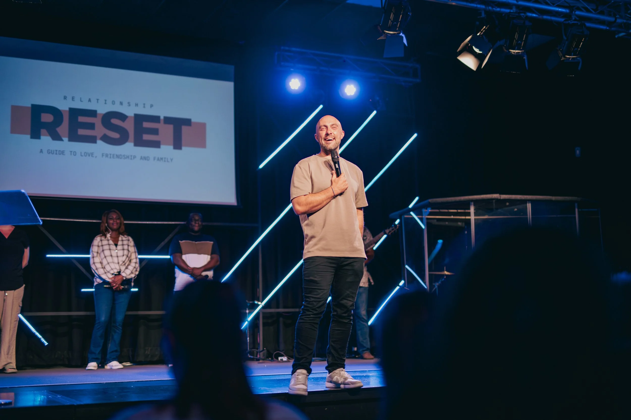 A man holding a microphone speaks on stage during a presentation or event, with a large screen behind him displaying the word "RESET" and the subtitle "A guide to love, friendship and family." There are a few other people on stage with him, and the stage is decorated with modern blue lights and diagonal light strips.
