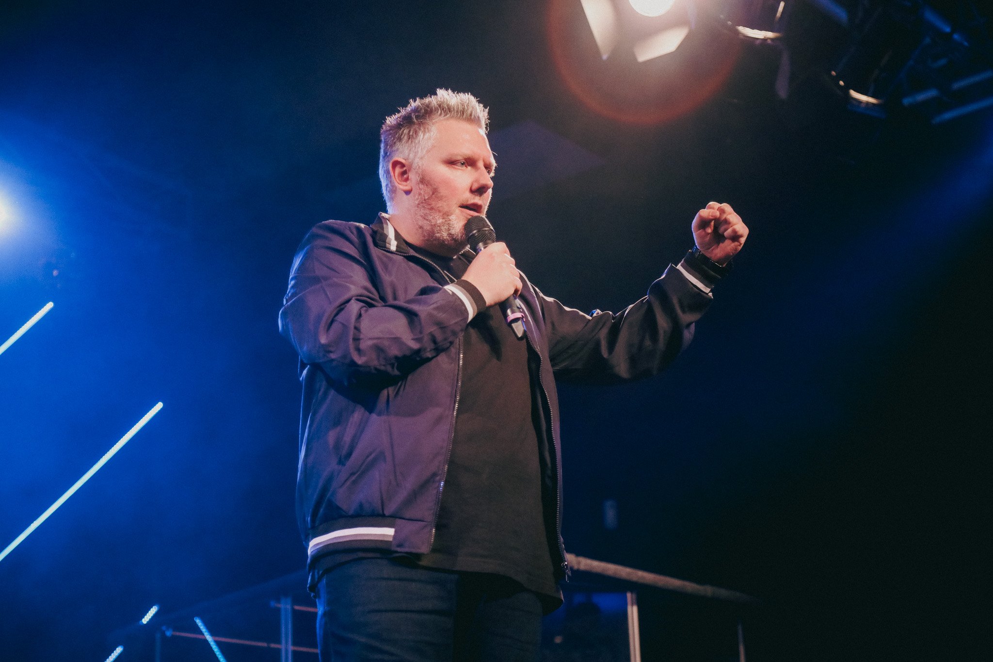 A man with gray hair and beard wearing a dark jacket speaking into a microphone on stage under blue and purple lighting.