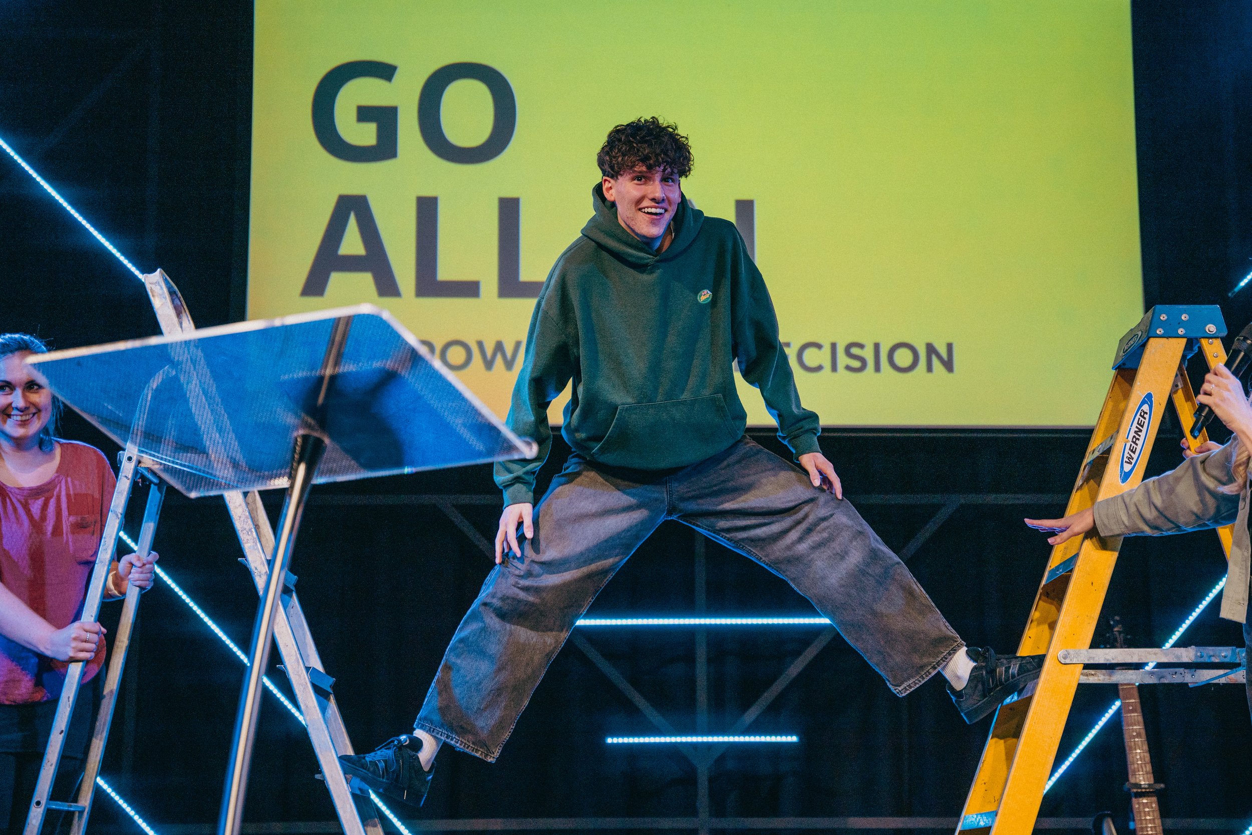 A young man is standing on a ladder with his arms and legs spread out wide, with a smile on his face. Two women are nearby, one holding a small ladder and the other speaking into a microphone. A large yellow screen in the background displays the words 'GO ALL IN' and smaller text that is partially visible.