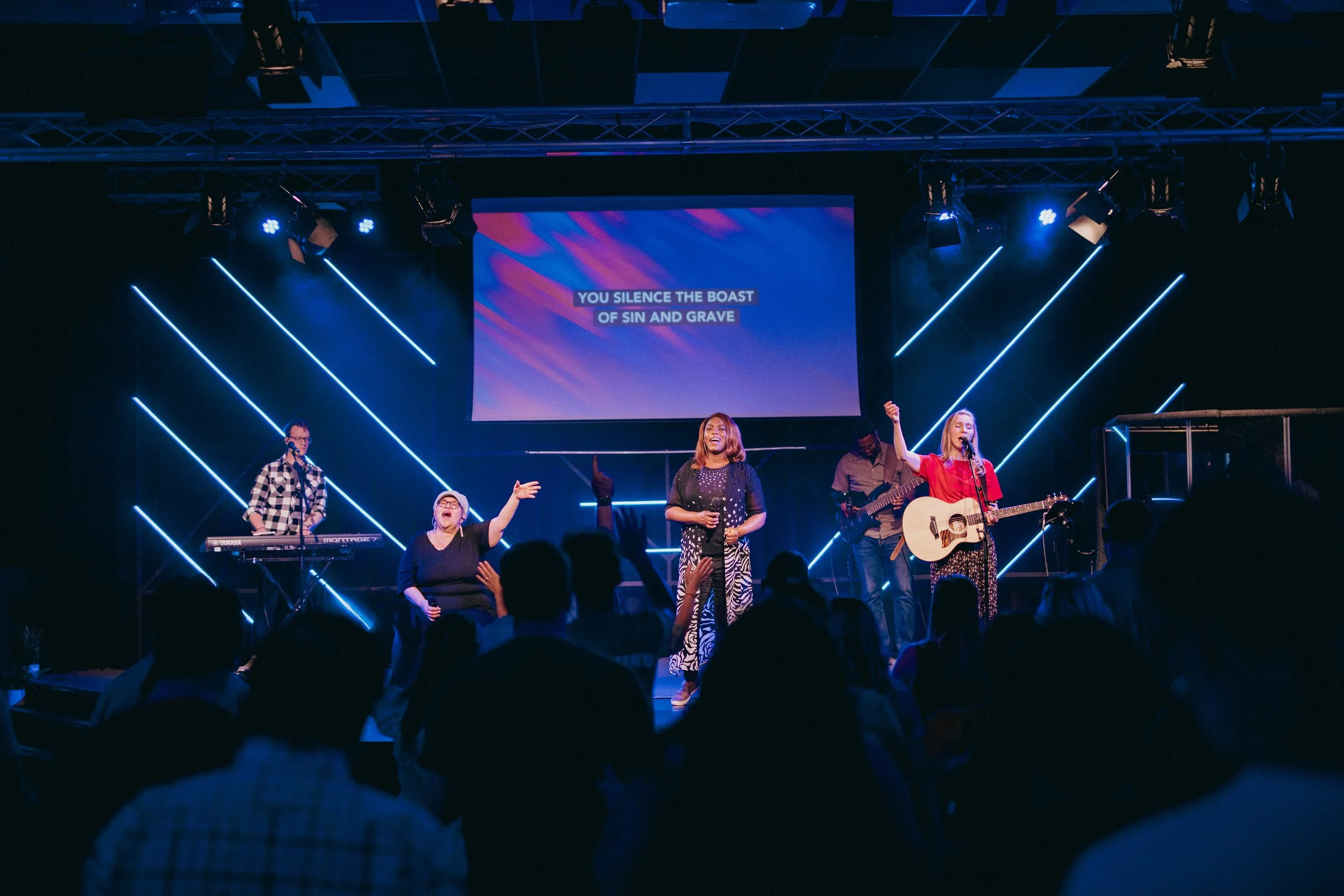 A live music performance on stage with four musicians, including two women singing and playing guitar, one woman leading with a microphone, and a man playing keyboard. The background has neon light designs and a large screen displaying the lyrics, 'YOU SILENCE THE BOAST OF SIN AND GRAVE.' The audience is visible in the foreground.