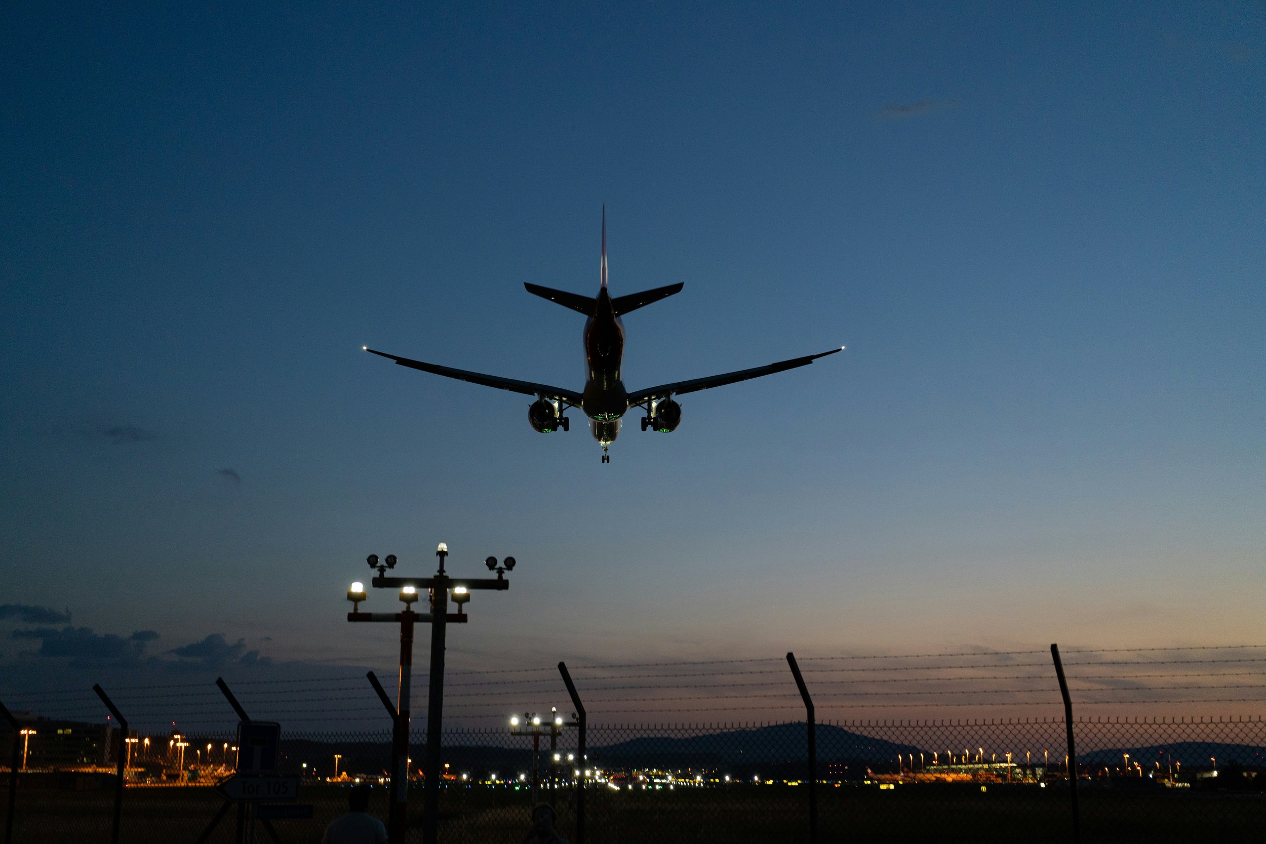 An airplane flying low over an airport runway during dusk with airport lights and runway fences visible.