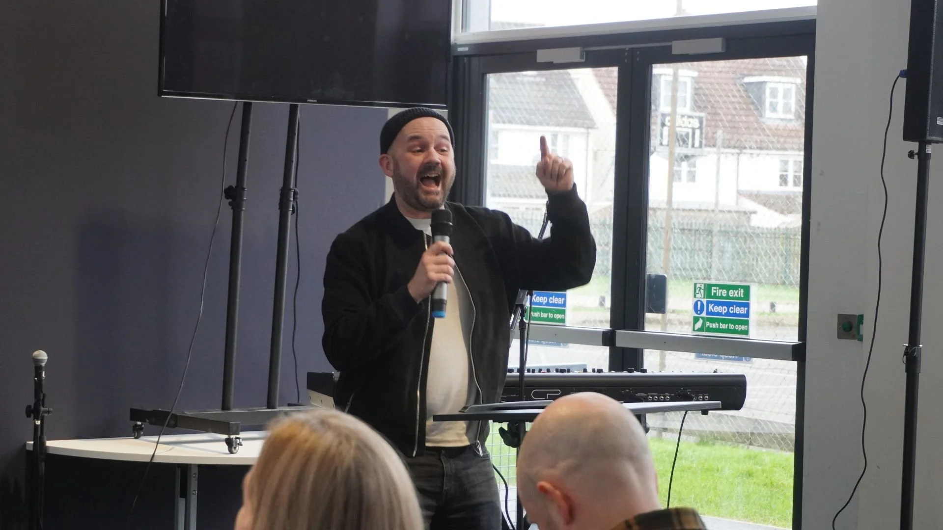 A group of people participating in a karaoke or singing event indoors. Some are holding microphones and singing, while others are dancing and enjoying the performance. A large screen displays lyrics in the background.