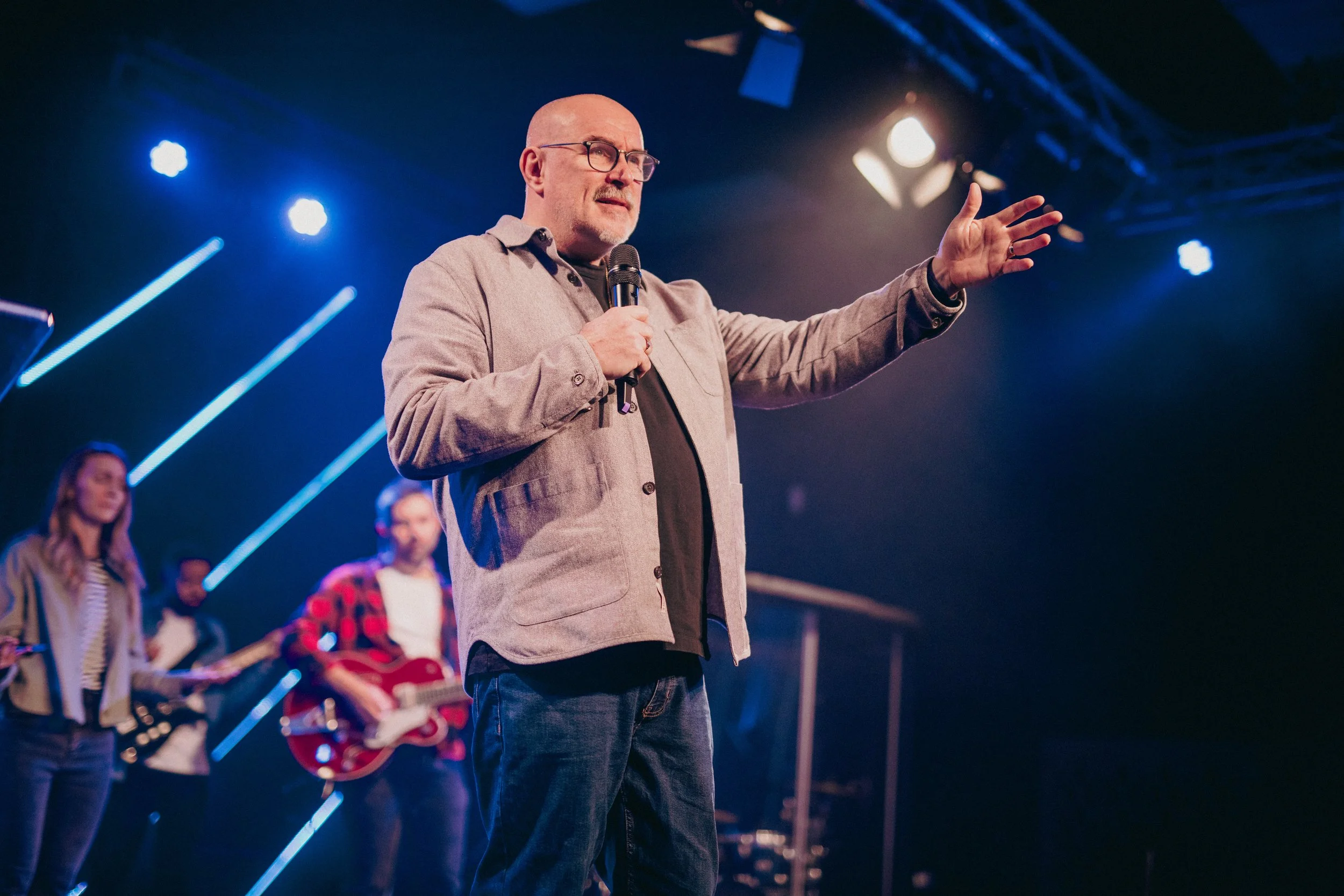 A man with glasses and a bald head holding a microphone on stage, gesturing with his right hand, with a band playing in the background under stage lights.