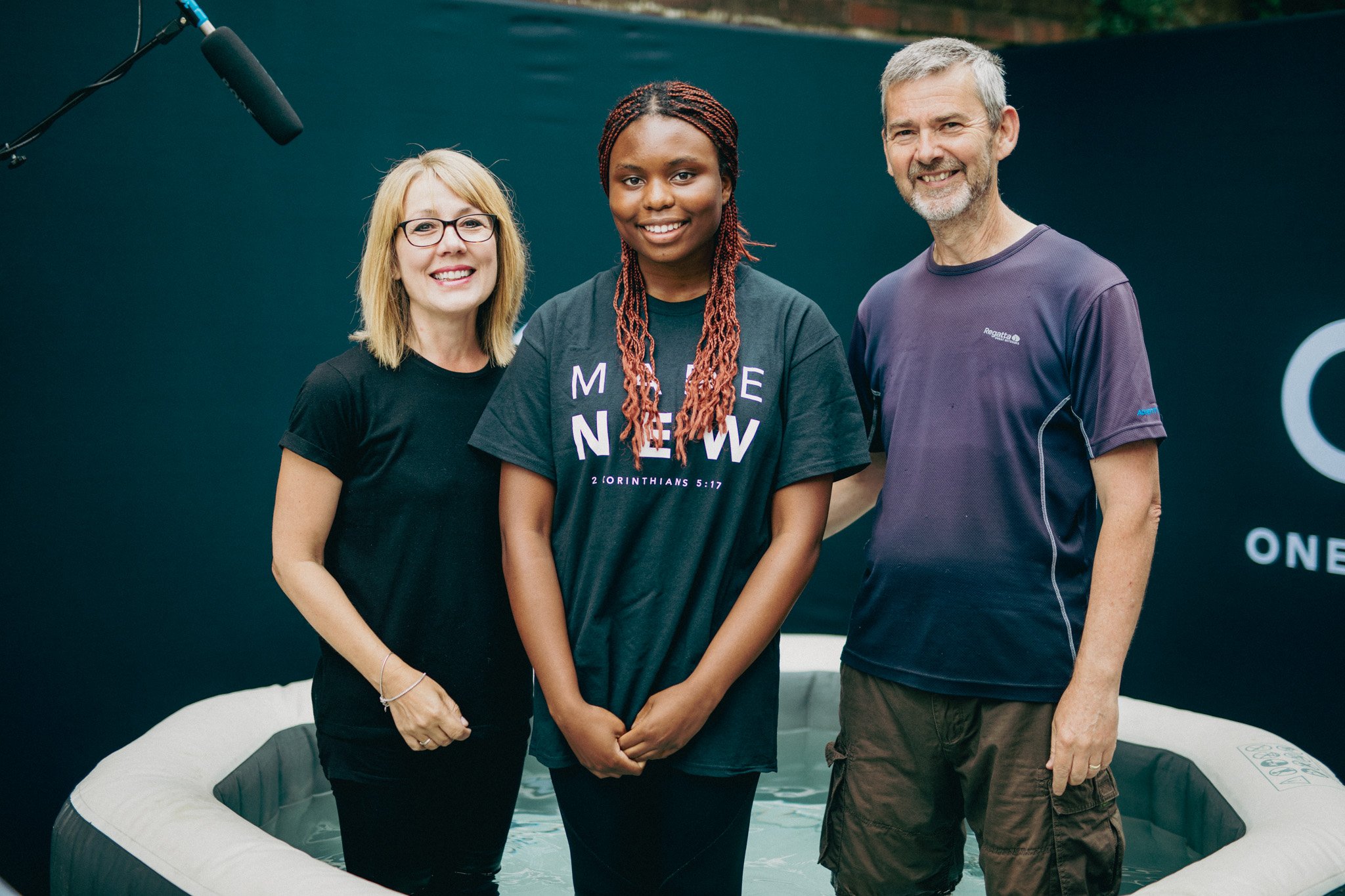 Three people standing outdoors, smiling, with a black backdrop, and a partially visible inflatable hot tub in front of them.