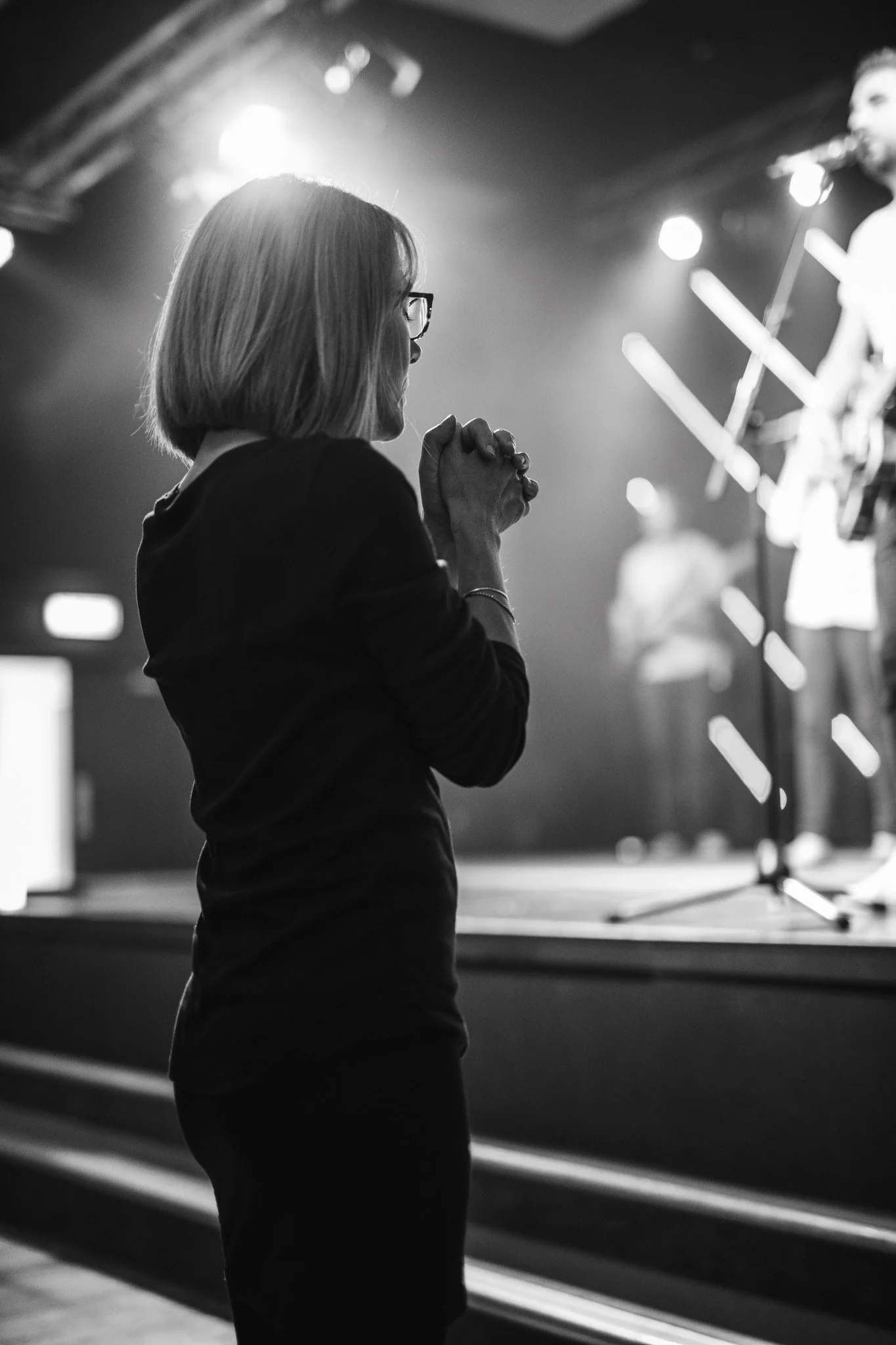 A woman with glasses and a short bob haircut, dressed in dark clothing, is standing with her hands clasped together in prayer or reflection, facing a stage with a musician performing with a guitar and microphone, illuminated by stage lights.