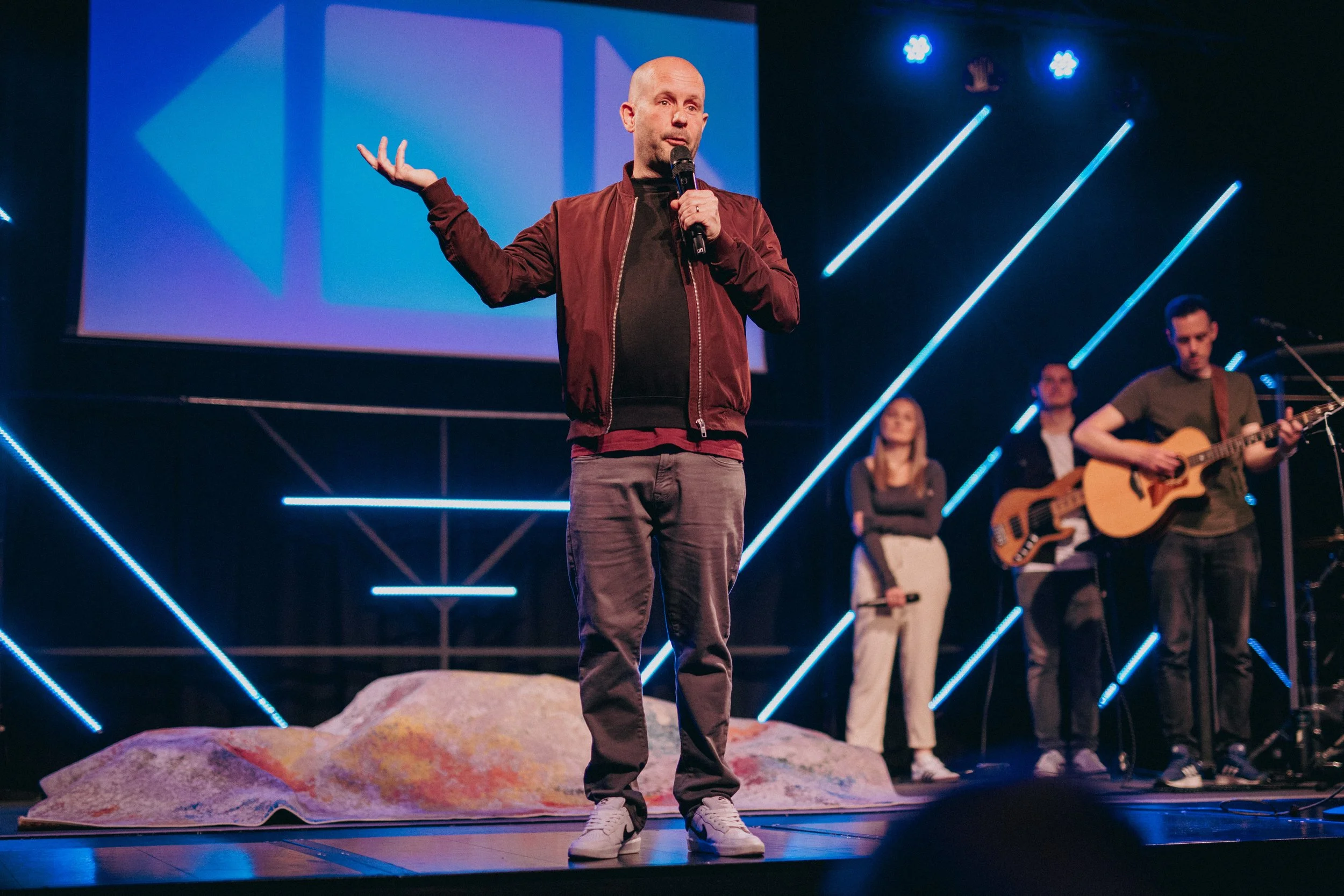 A man in a maroon jacket and gray pants is speaking into a microphone on a stage with a colorful background and three band members playing guitars and bass in the background.