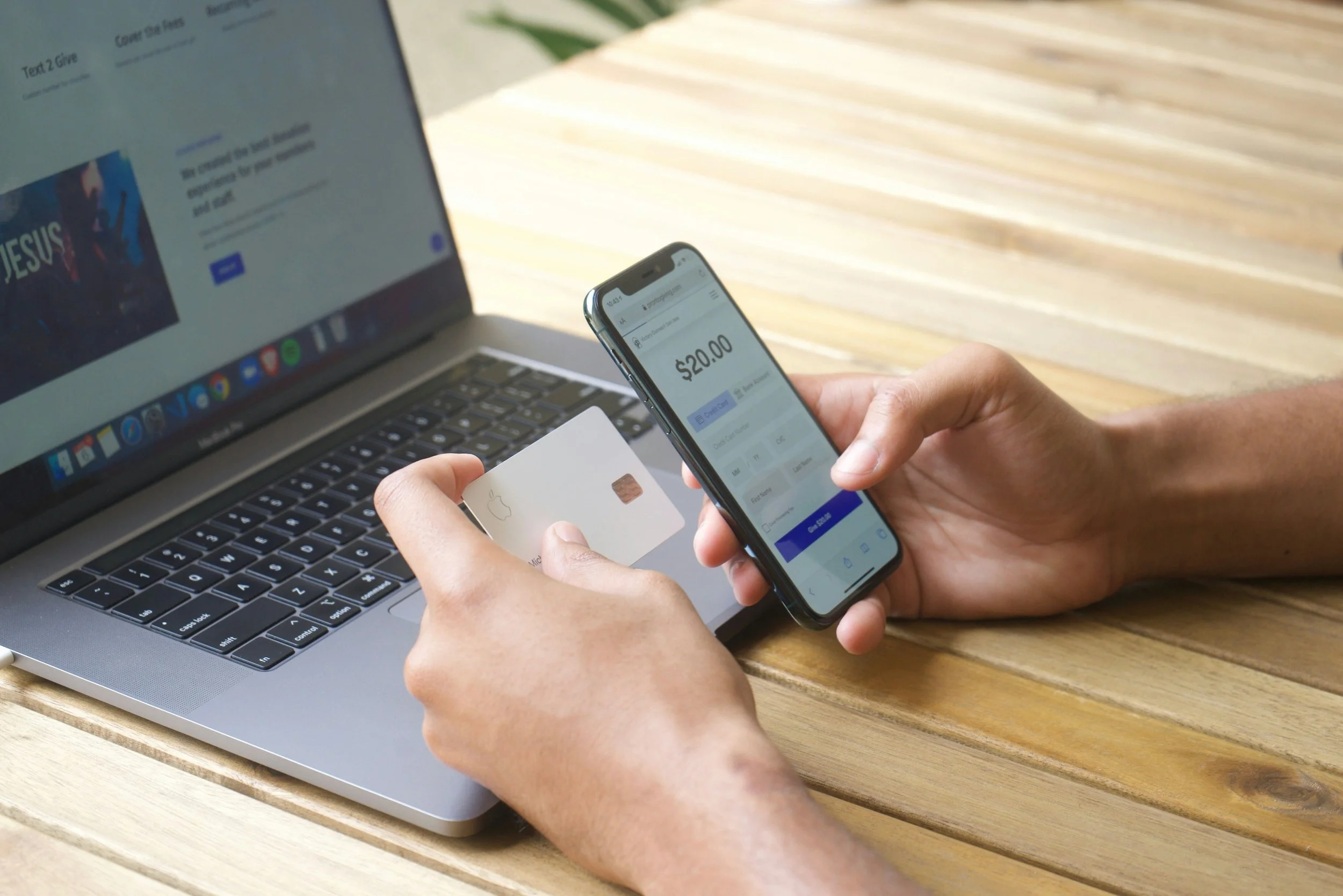 Person making an online payment with a smartphone at a wooden desk, using a credit card, laptop in the background, showing a $20.00 transaction on the phone screen.