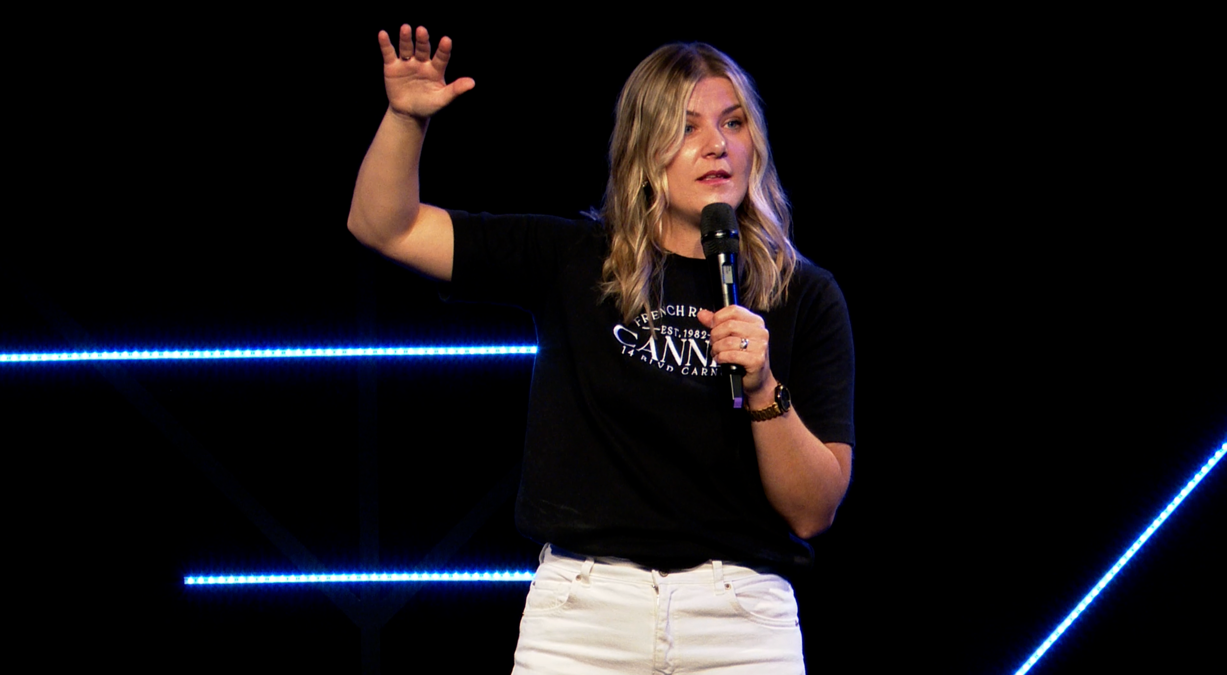 A woman with blonde hair holding a microphone and gesturing with her right hand while speaking on stage with black background and blue LED lights.