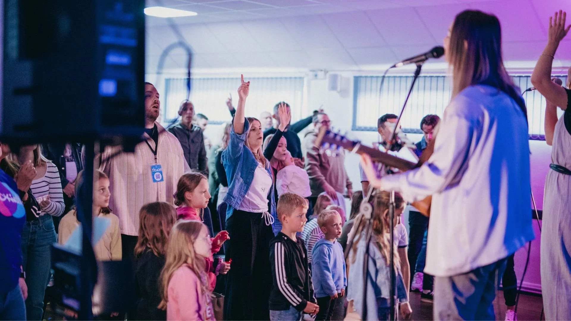 People attending a church service or event in a room with a small stage and a screen displaying worship lyrics.