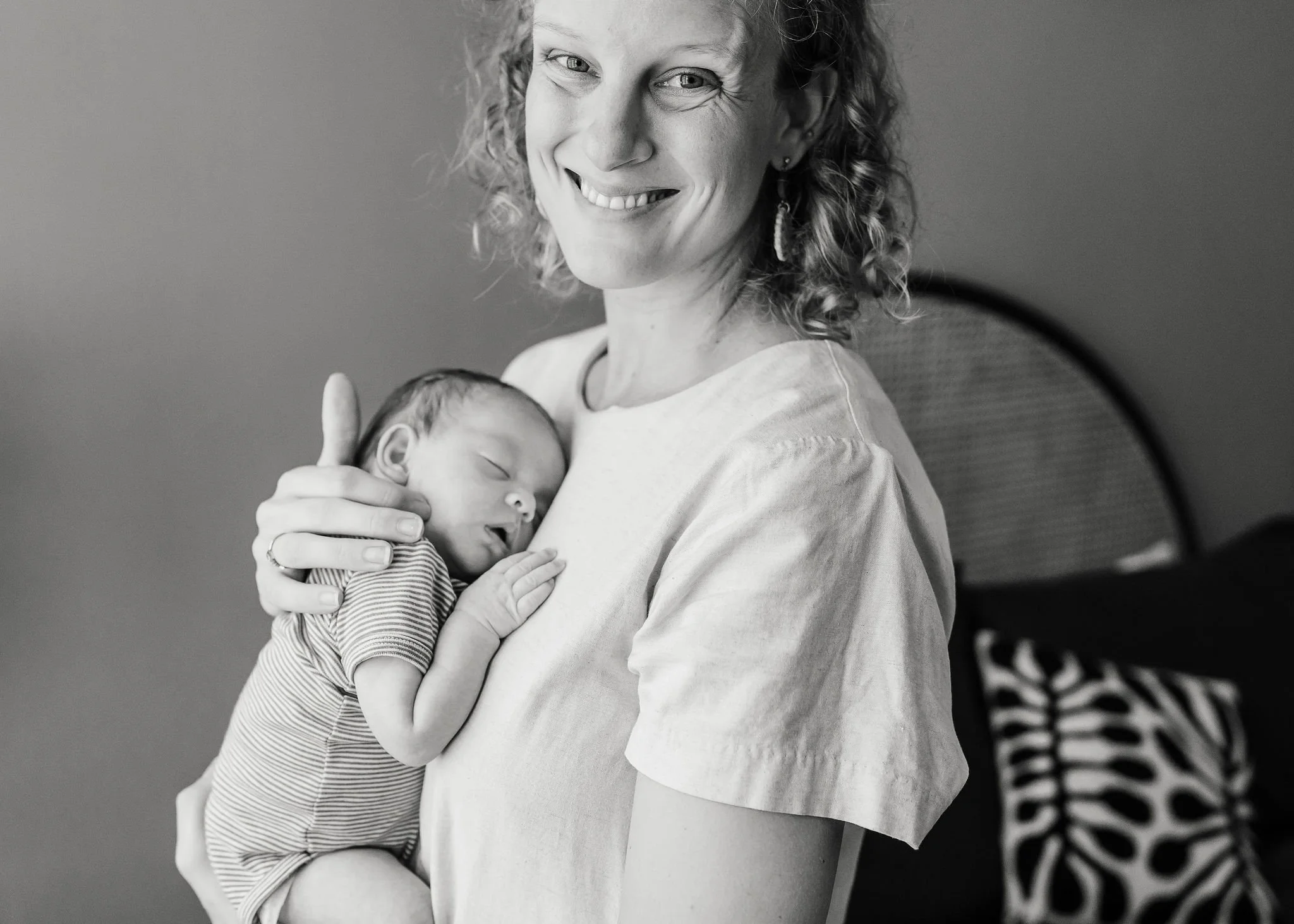 A woman with curly hair smiling while holding a sleeping newborn baby close to her chest.