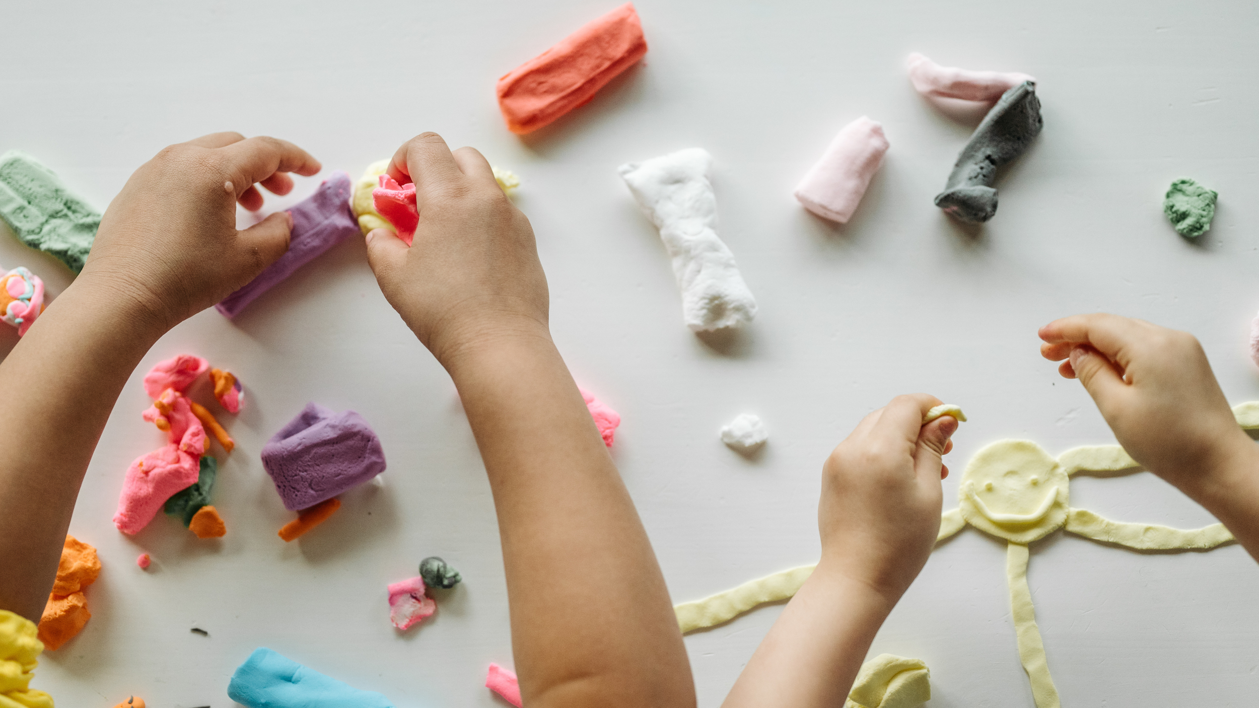 Two children playing with colorful play dough on a white surface, with a sun drawn out of yellow dough.